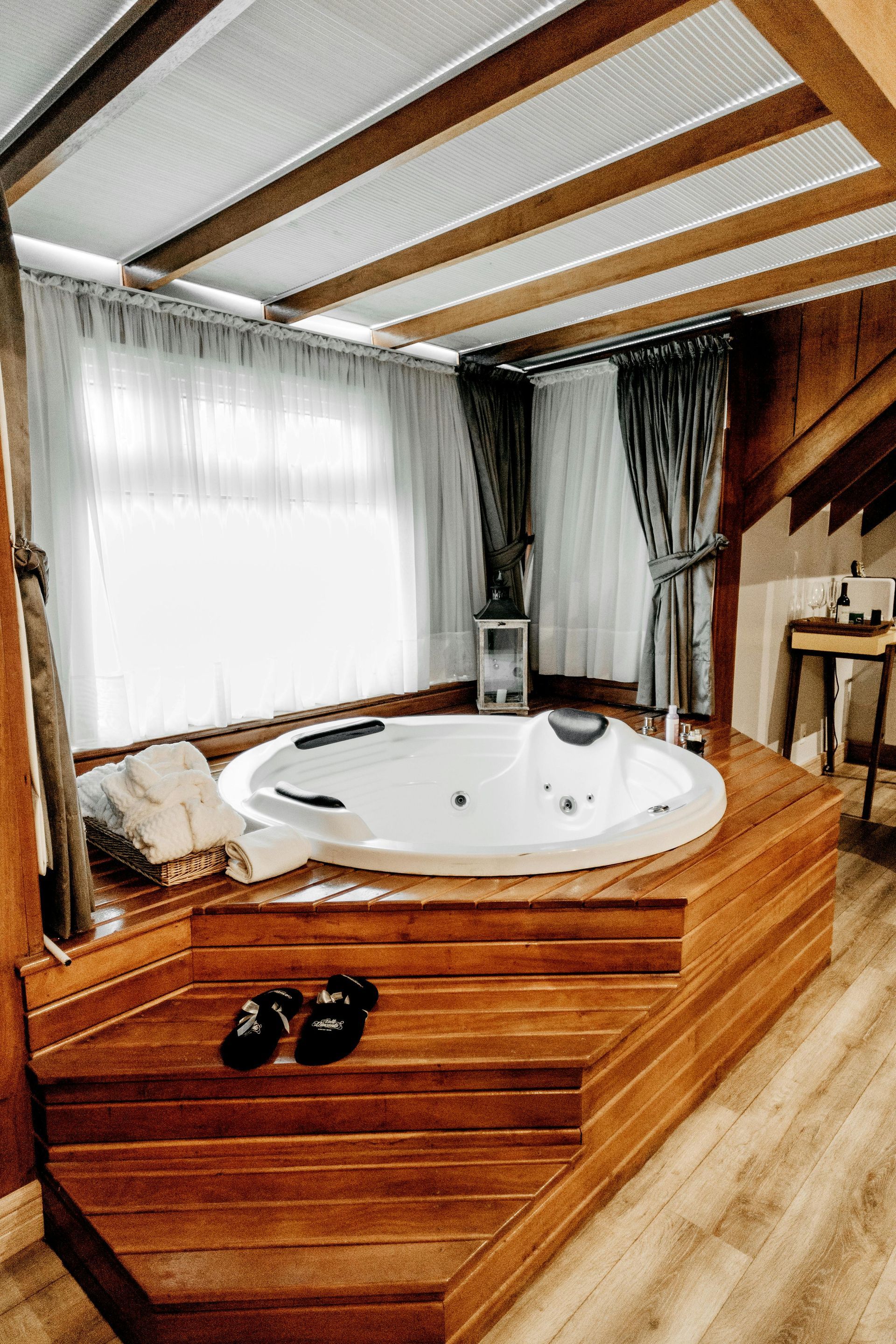 Indoor hot tub on a wooden platform near a window with white curtains; brown wooden ceiling.