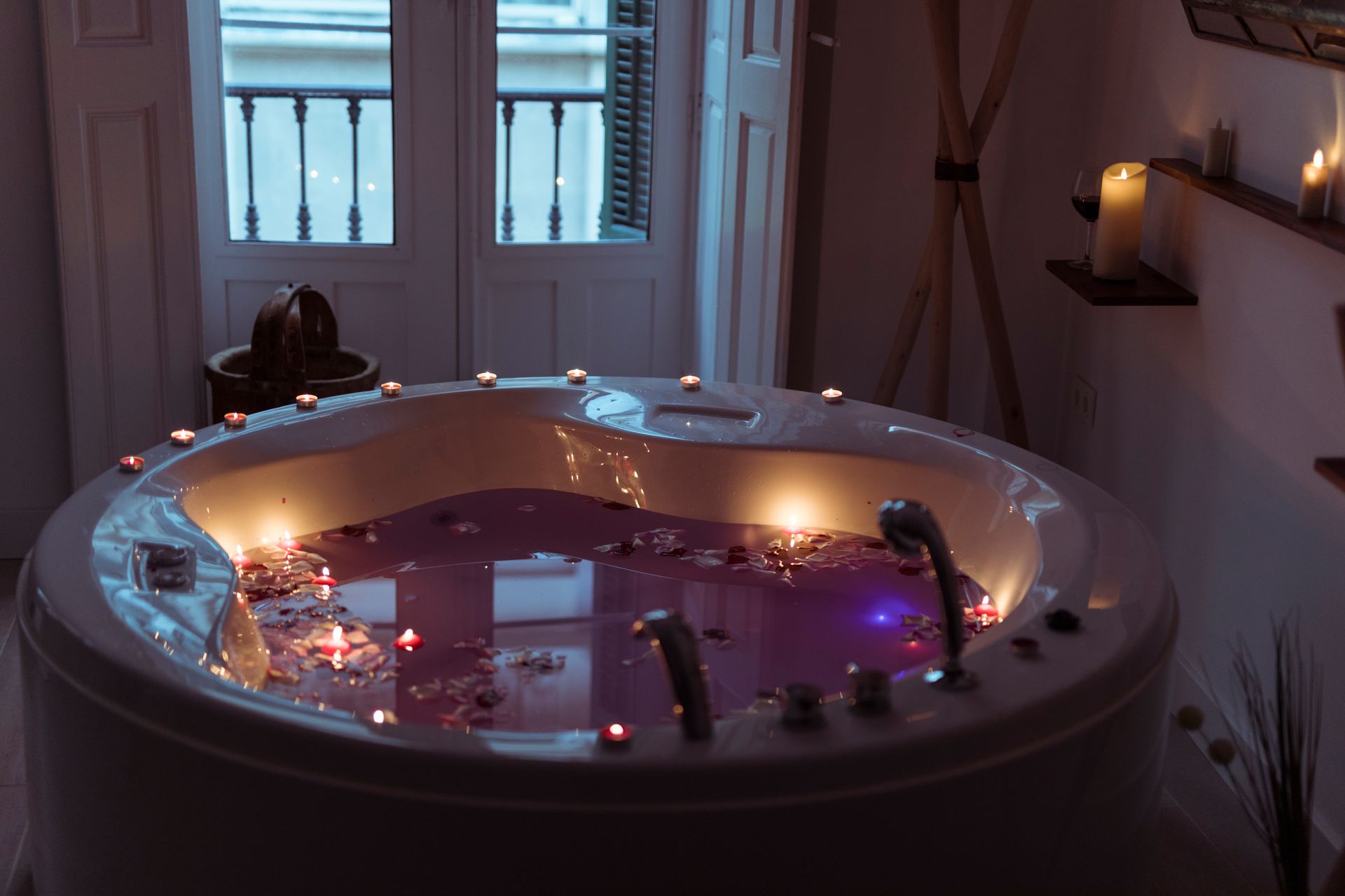 Round bathtub with pink water, candles, and rose petals in a dimly lit spa-like room.