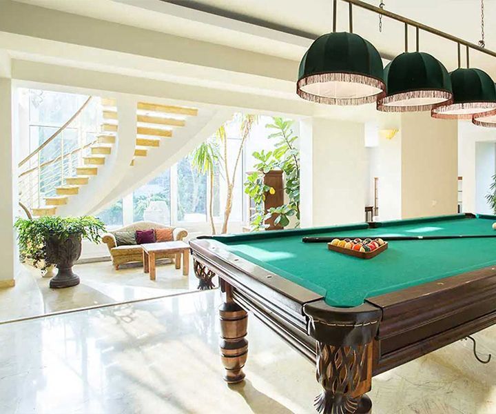 Pool table in a bright room with a view of a staircase, sofa, and outdoor plants. Green felt, dark wood.