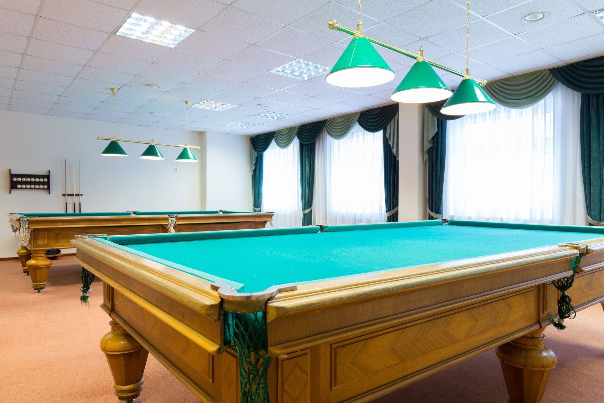 Two pool tables in a room with green felt, green lamps, and light-colored wood.