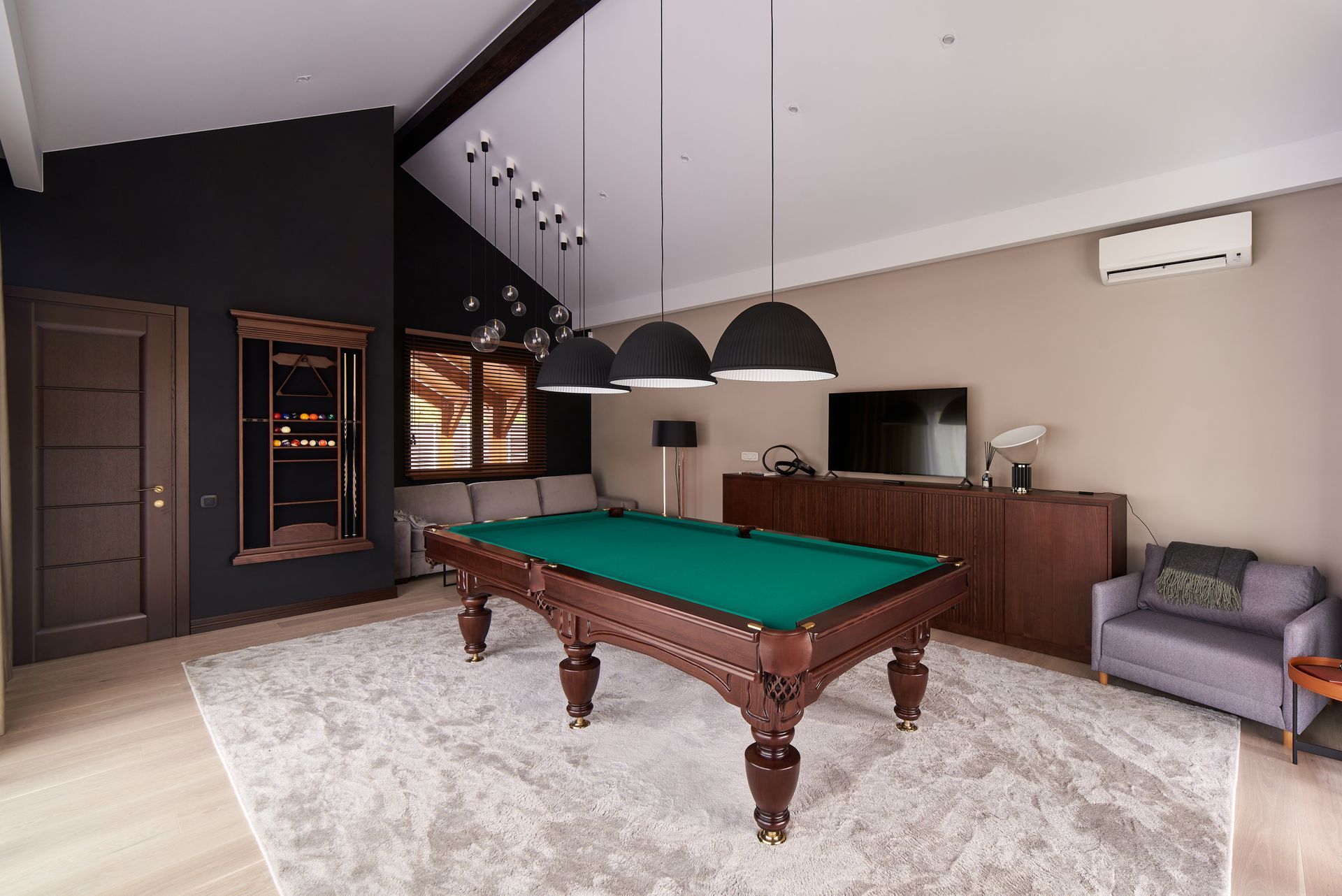 Pool table in a modern game room, with dark walls, pendant lights, and a TV.