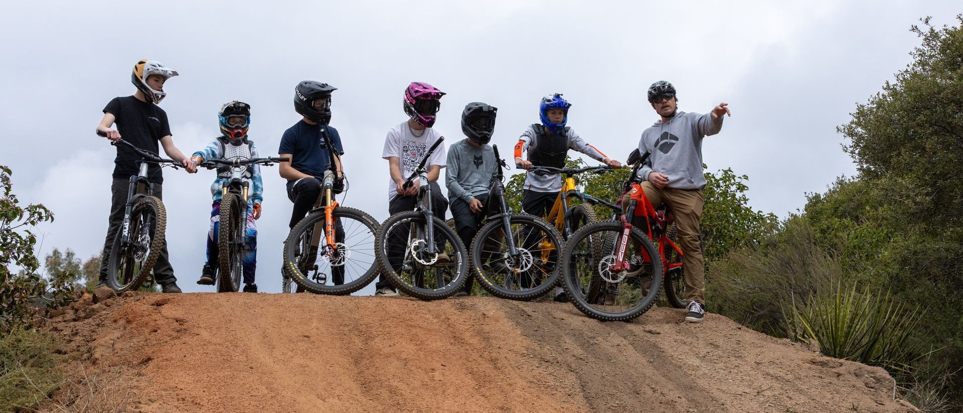 A group of mountain bikers, wearing helmets, stand on a dirt mound with their bikes, under an overcast sky.