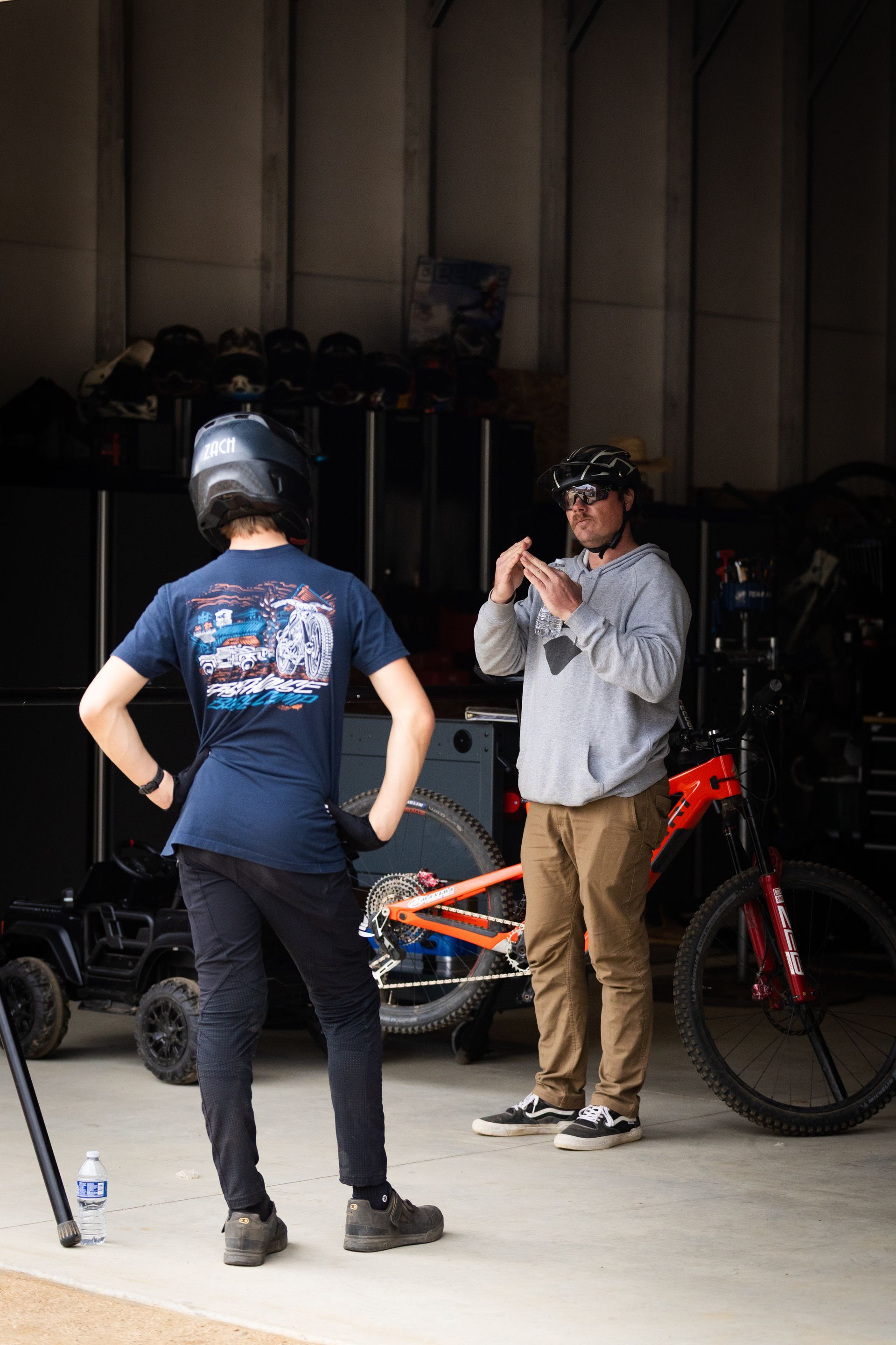 Two men in a garage; one wearing a helmet and gazing at the other who is holding a small object near an orange mountain bike.