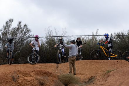 People on bikes at a dirt bike track. A man in a gray shirt gives instruction as bikers wearing helmets prepare to ride.