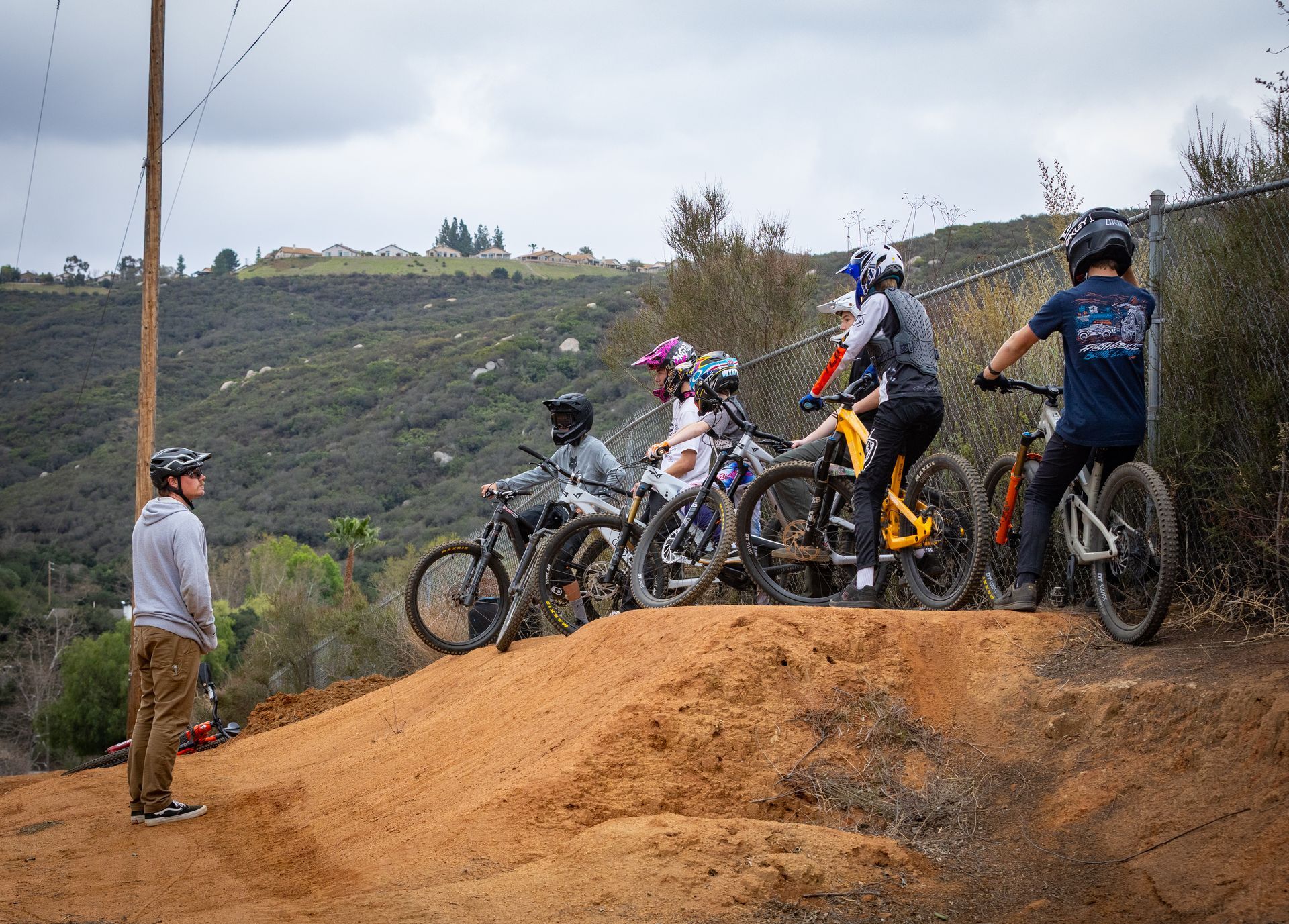 A group of kids on mountain bikes preparing to ride a dirt jump, watched by a coach. 