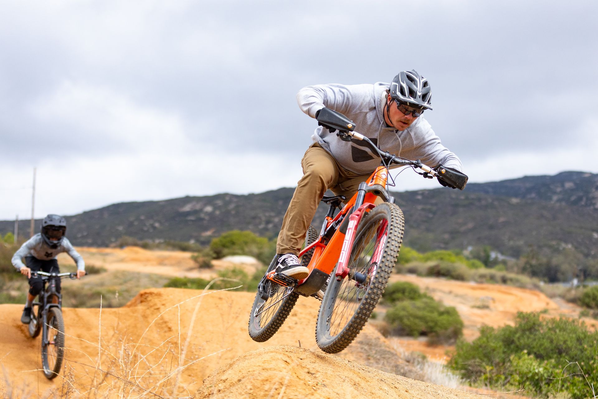 Two people on mountain bikes, one airborne on a dirt jump. 