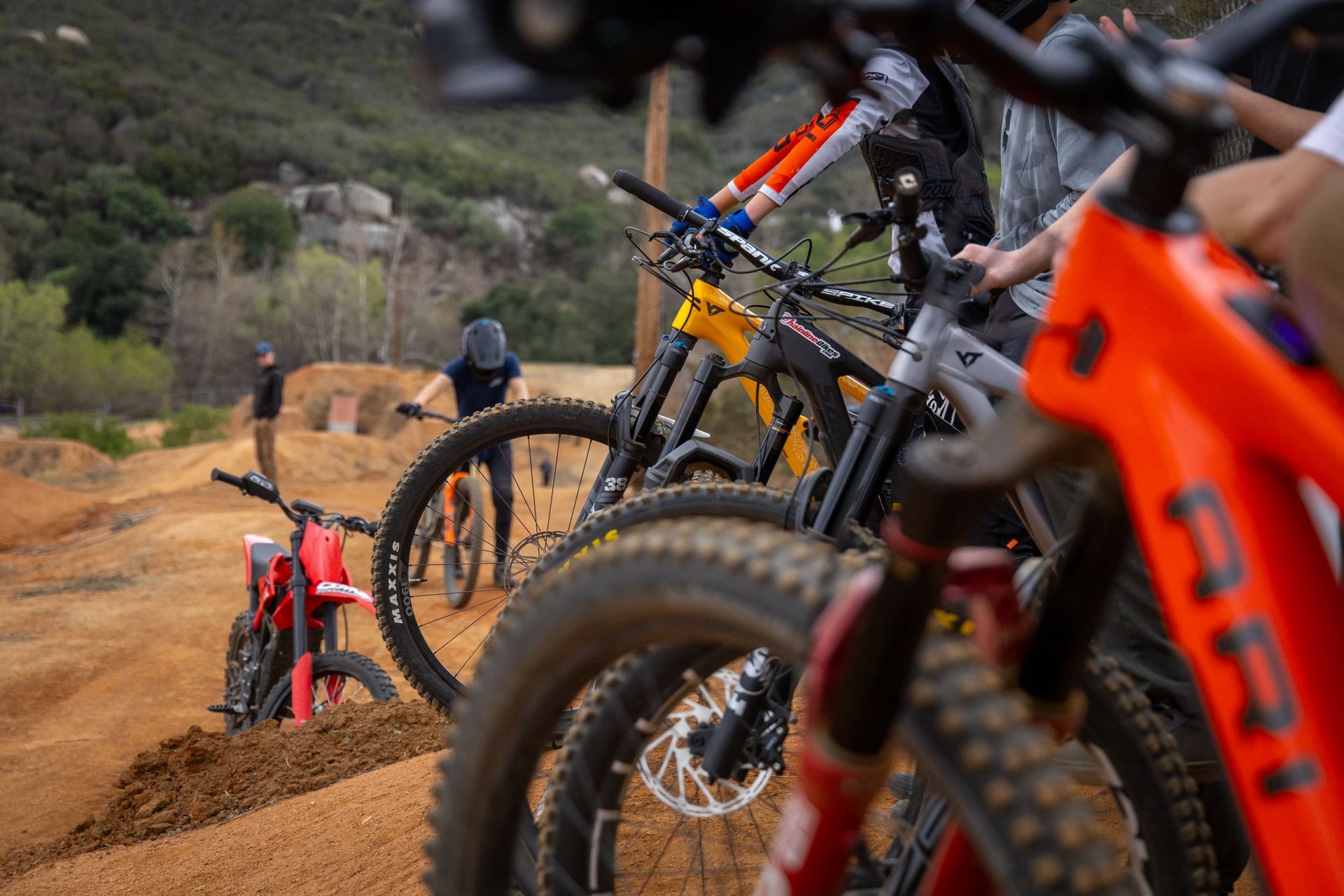 Mountain bikes lined up, riders preparing to start on a dirt track course. A person is riding in the distance.