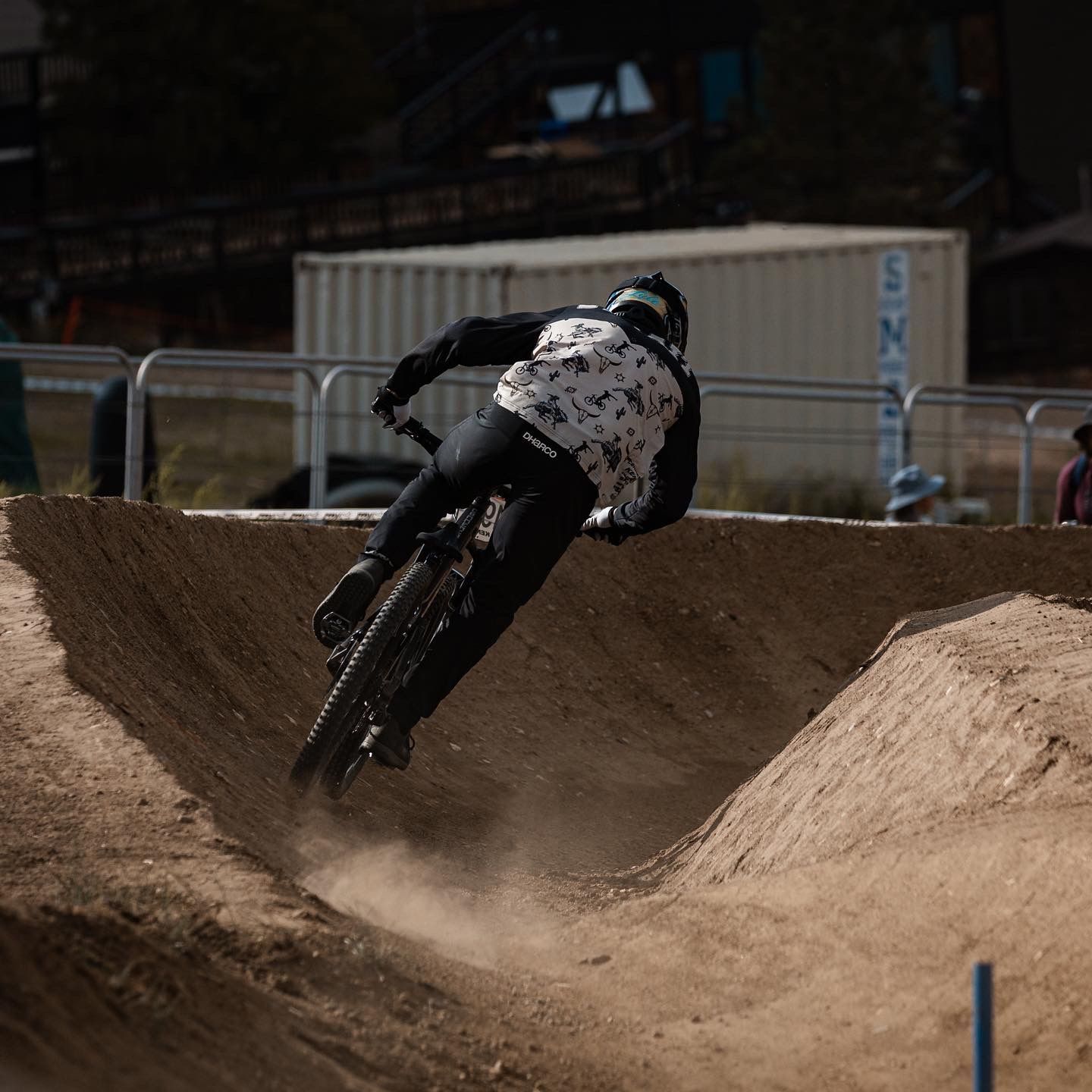 Mountain biker in a floral patterned jacket leans into a dirt berm on a course, kicking up dust.