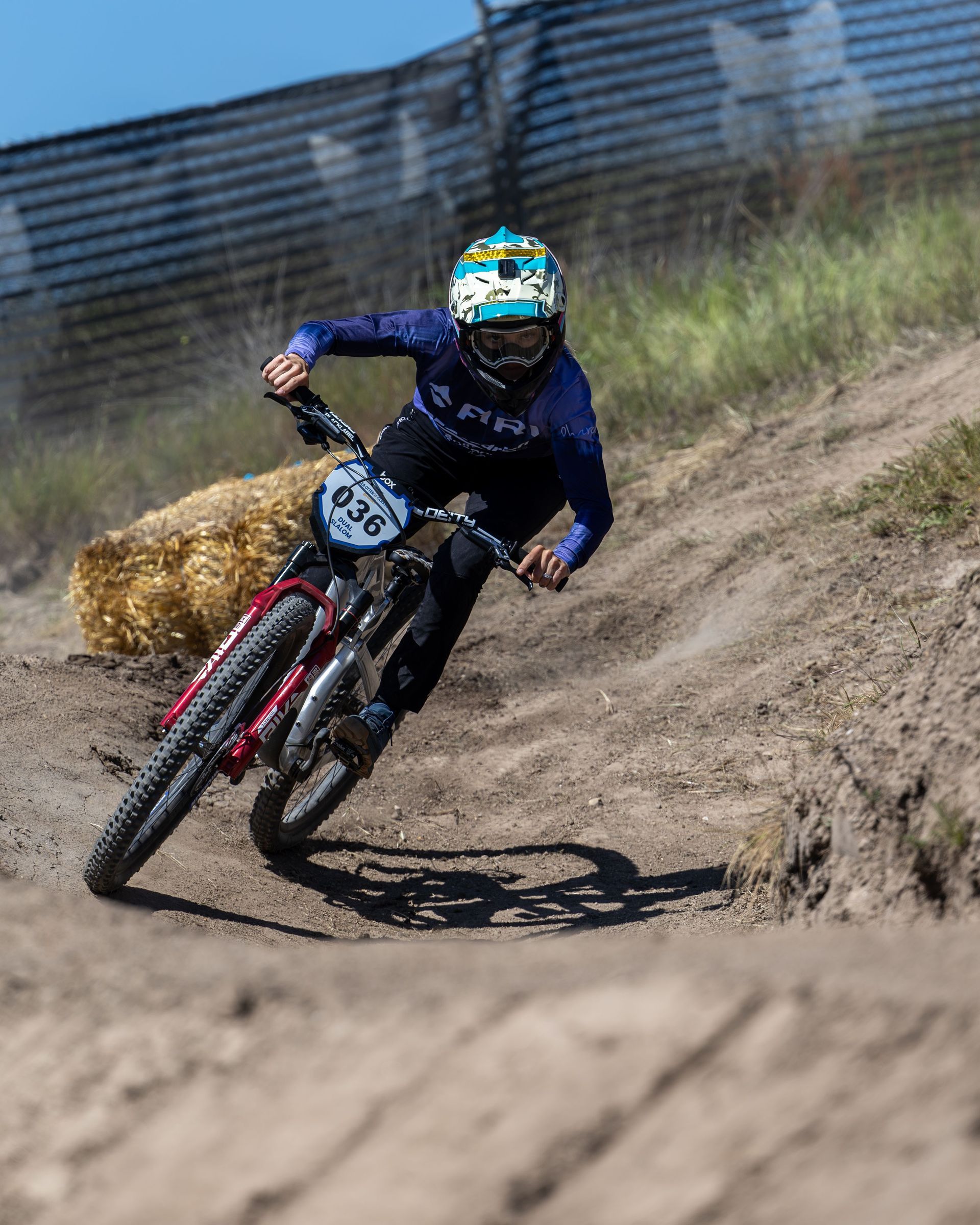 Mountain biker in blue gear leans into a turn on a dirt track, with a hay bale on the side.