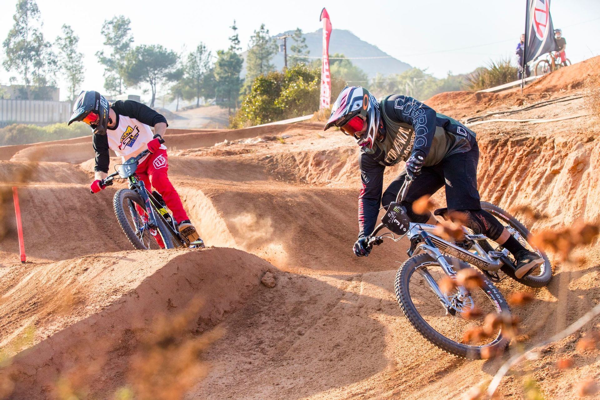 Two mountain bikers racing on a dirt track, kicking up dust. 