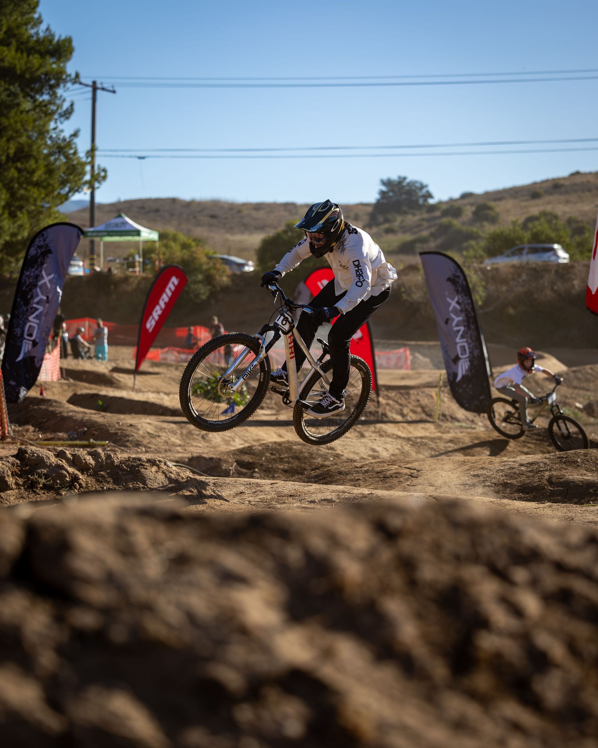 A mountain biker jumps over a dirt mound on a course. The rider wears a white and black outfit. 
