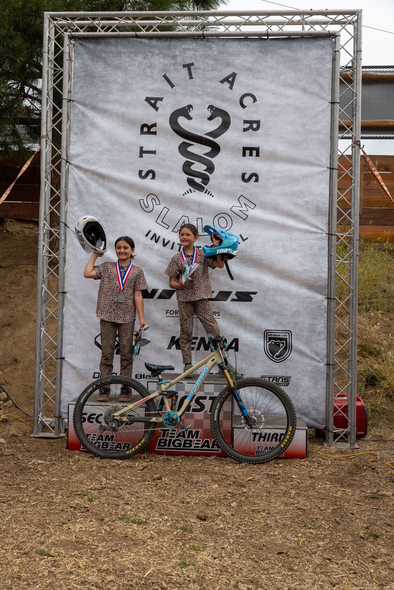 Two young mountain bikers on a podium, wearing medals and holding helmets, with a bike in front of a 