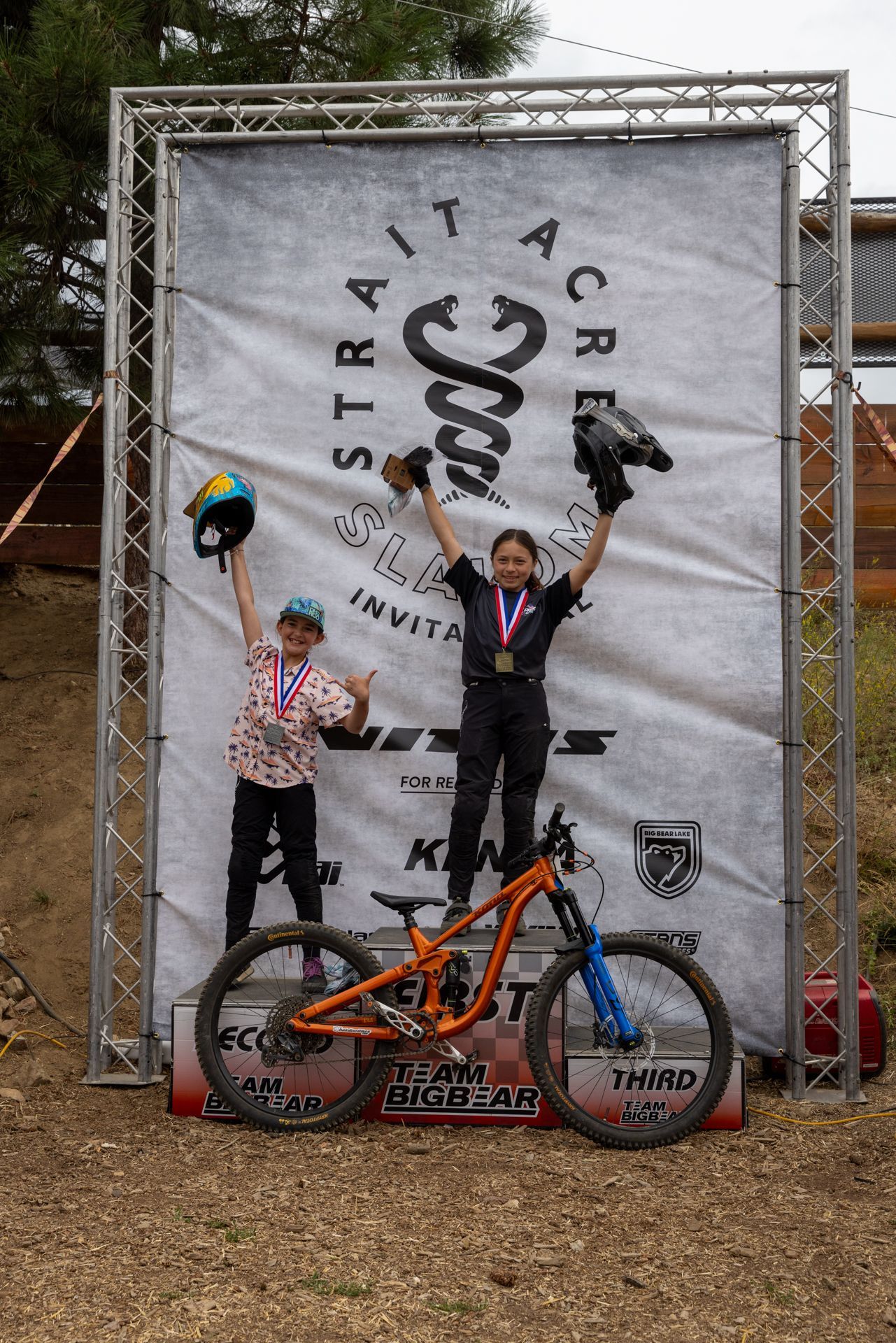 Two people on a podium celebrate a mountain biking win. One raises a helmet, the other raises gloves, both with medals.