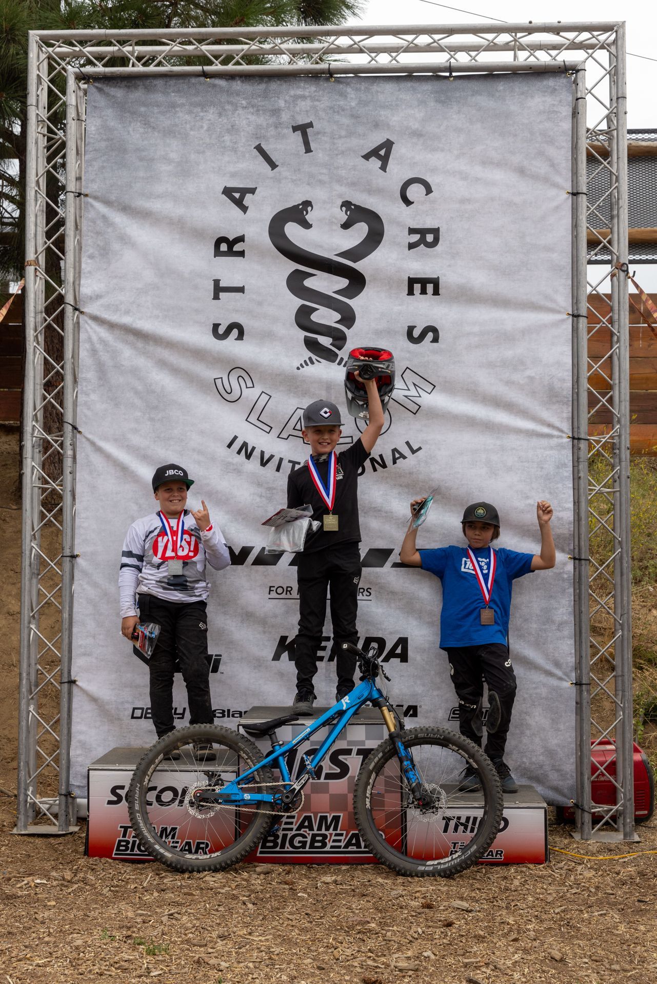 Mountain bike race winners on a podium. A boy in a black shirt raises his helmet, flanked by boys with medals,