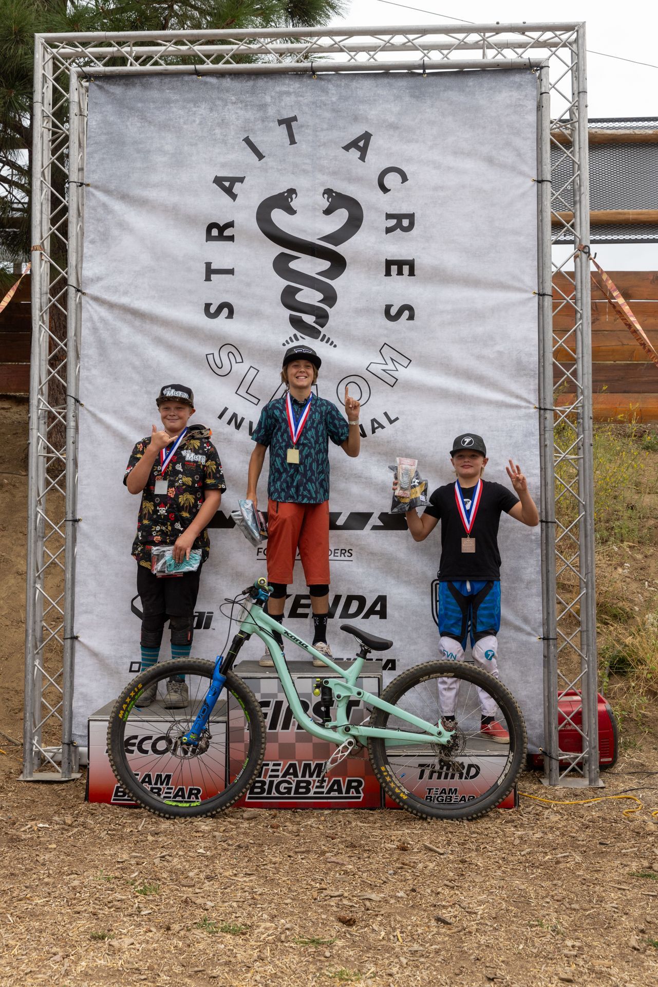 Three young mountain bikers on a podium, celebrating a race. The winner holds a trophy and flashes a peace sign. 