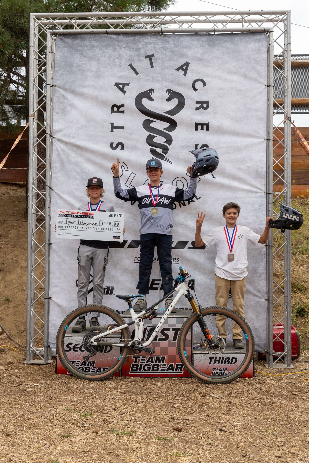 Podium with mountain bikers at Strait Acres. Winner holds helmet, second place holds check, third place has a medal.