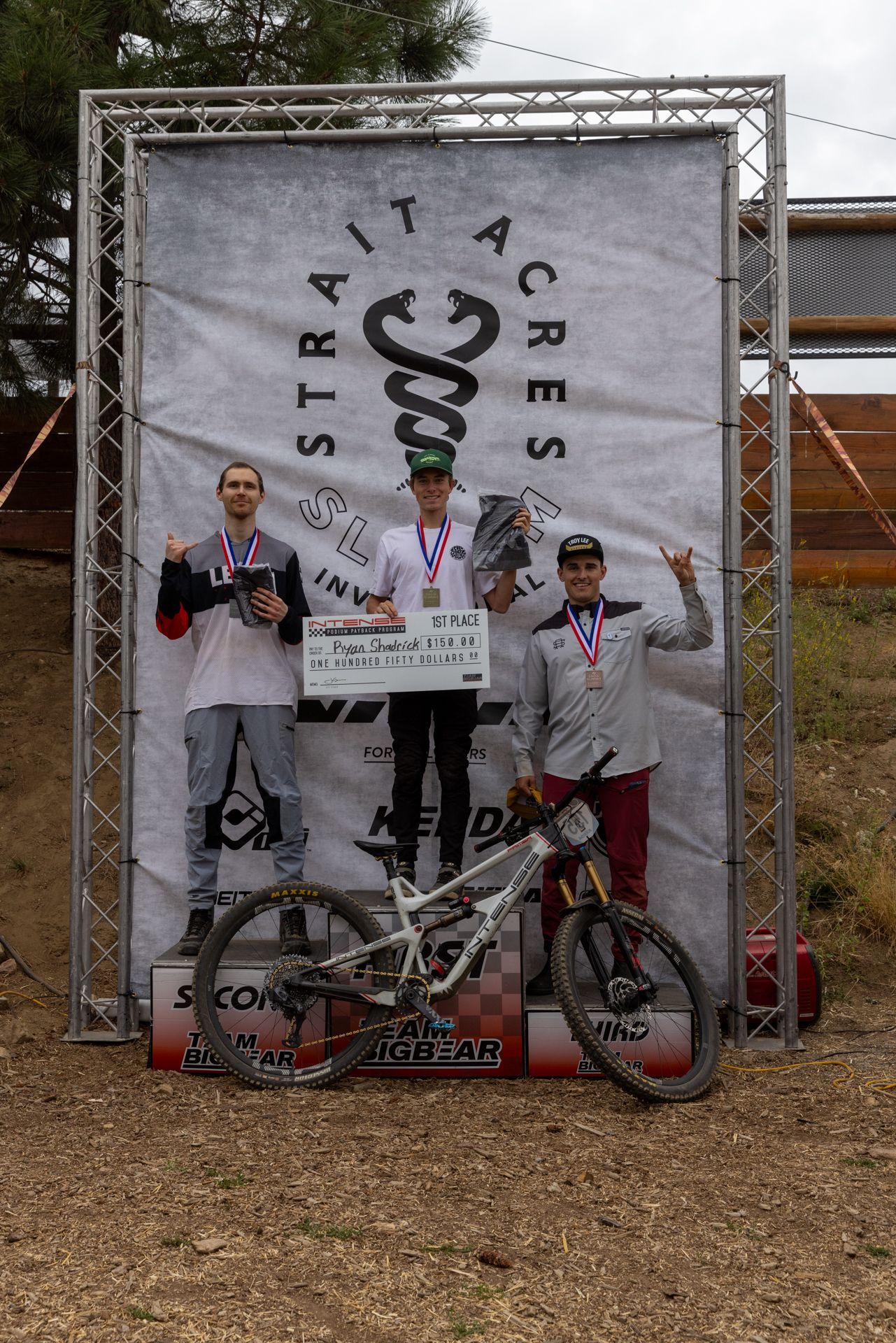 Podium with three mountain bikers at Strait Acres race. Winners hold medals and trophies, with a check displayed.