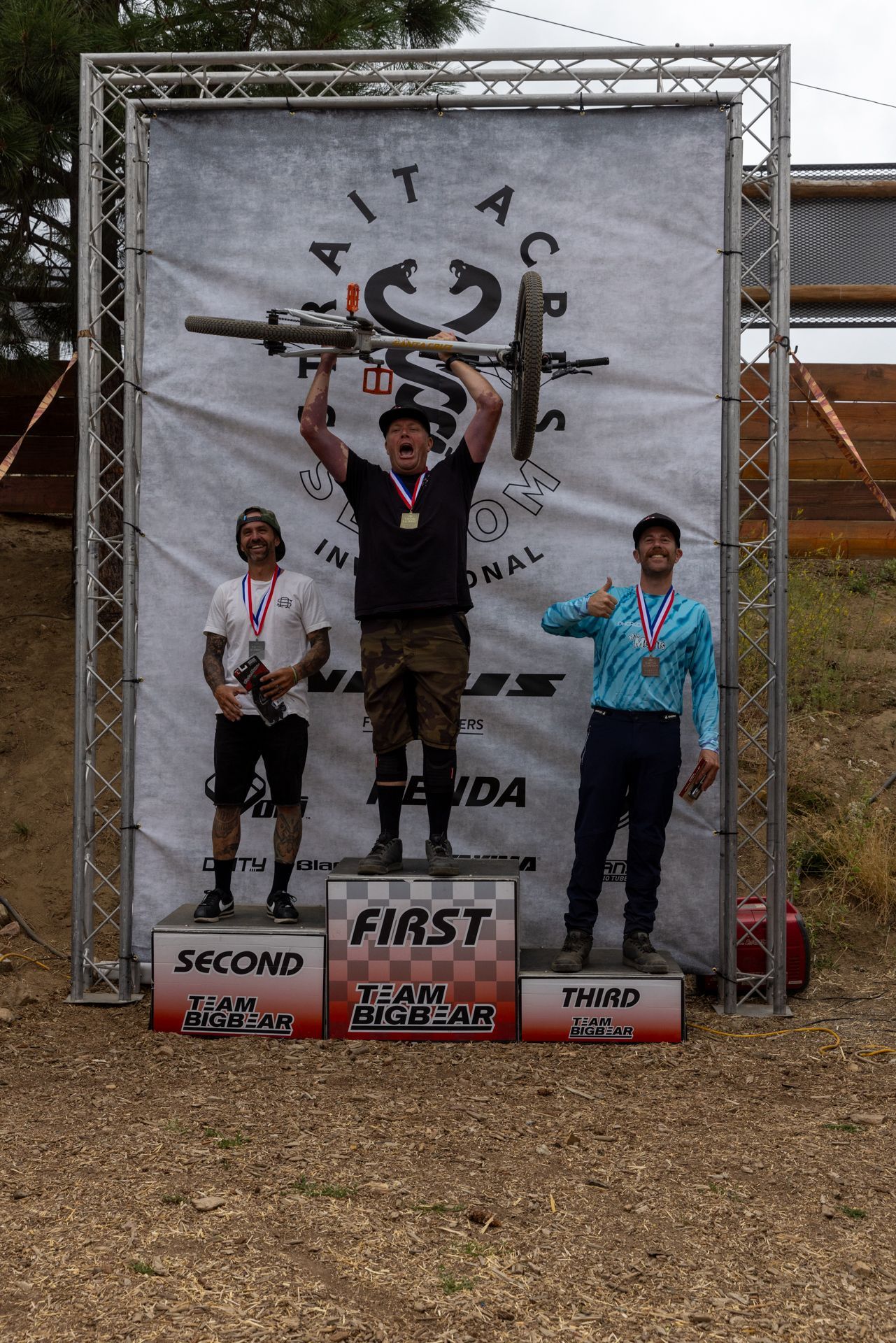 Mountain bike race podium: Winner raises bike, flanked by second and third place finishers, Nevada mountains in background.