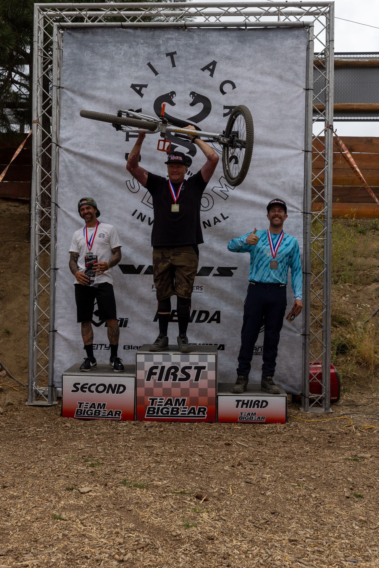 Podium with three mountain bikers: first place raises bike overhead, second holds trophy, third gives a thumbs-up