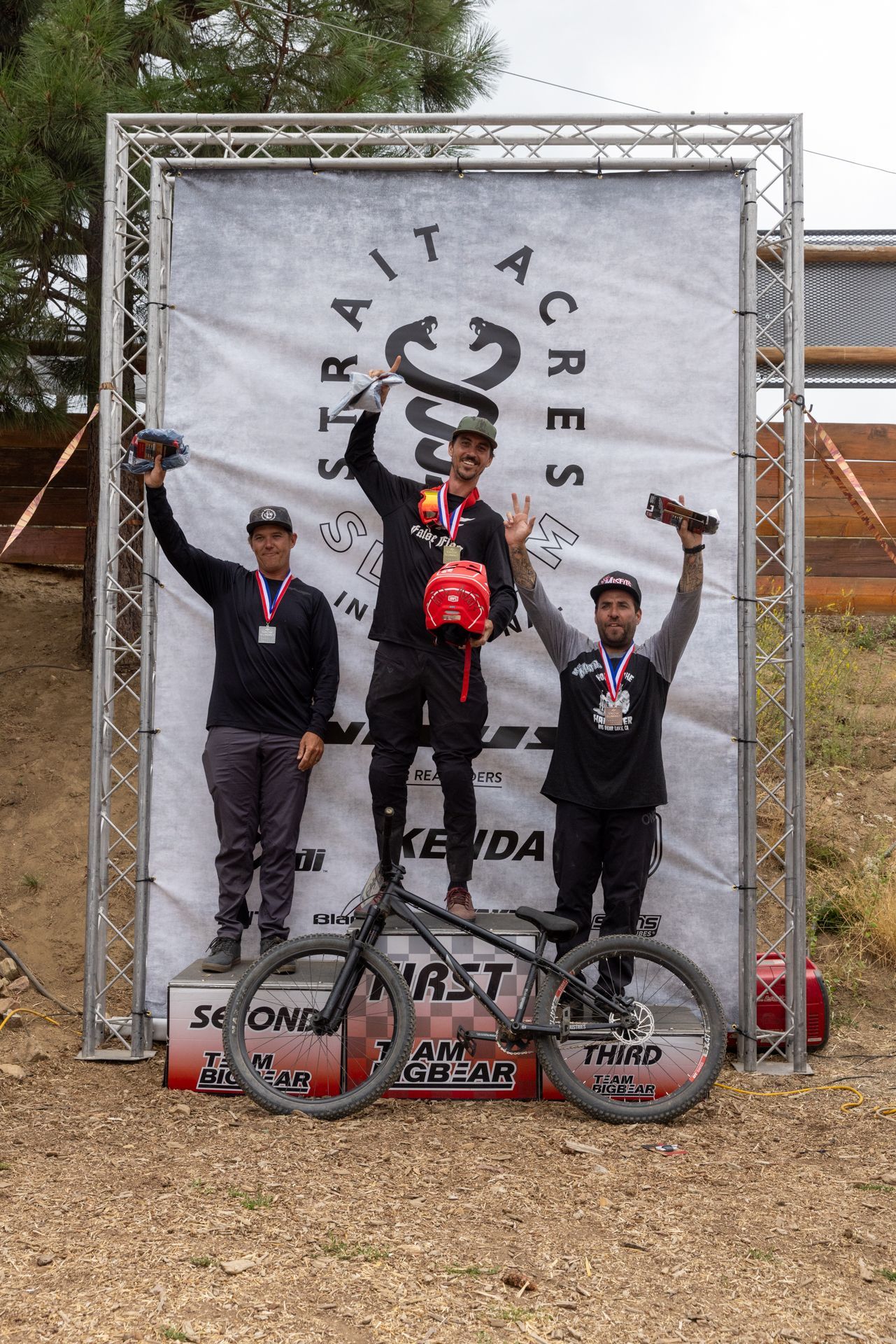 Winners of a bike race stand on a podium, arms raised. The event is at Spirit Acres, with a black bike in the foreground.