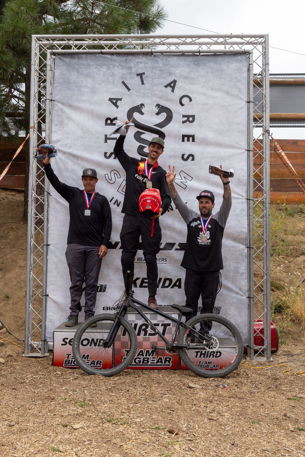 Podium of three cyclists: first place holds a trophy, second and third raise their hands. 