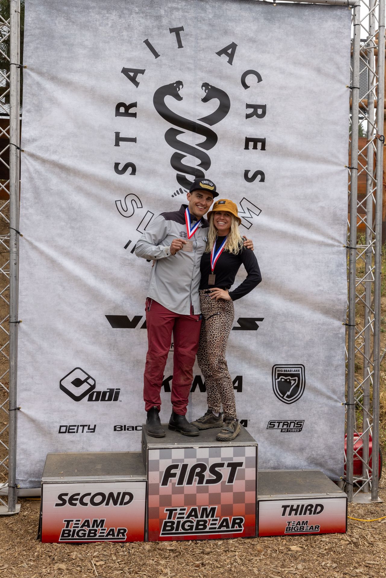Man and woman on podium, first place, smiling, medals. Banner: Strait Acres.