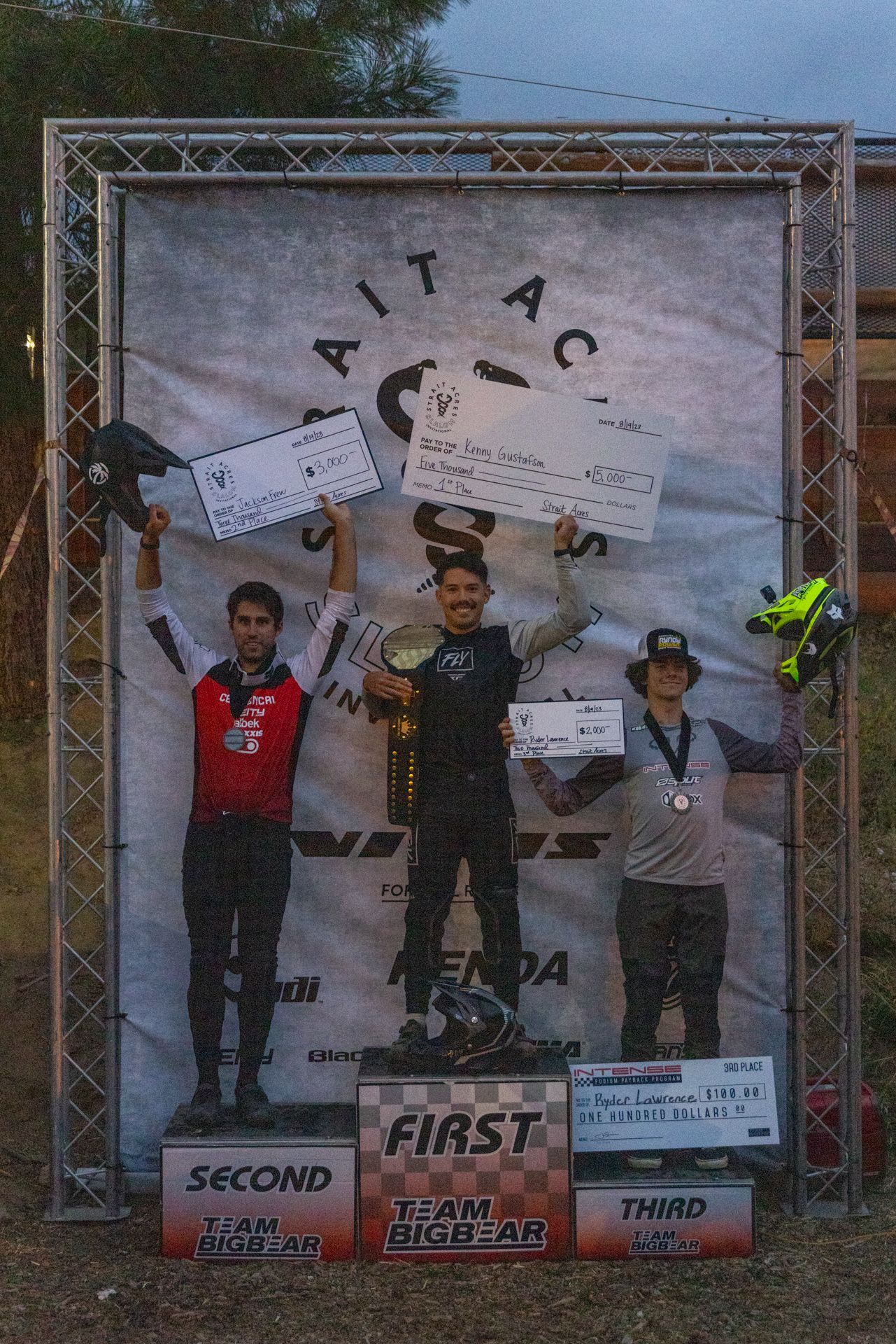 Podium with three men holding prizes. First place in the center, smiling, with a trophy and check. 