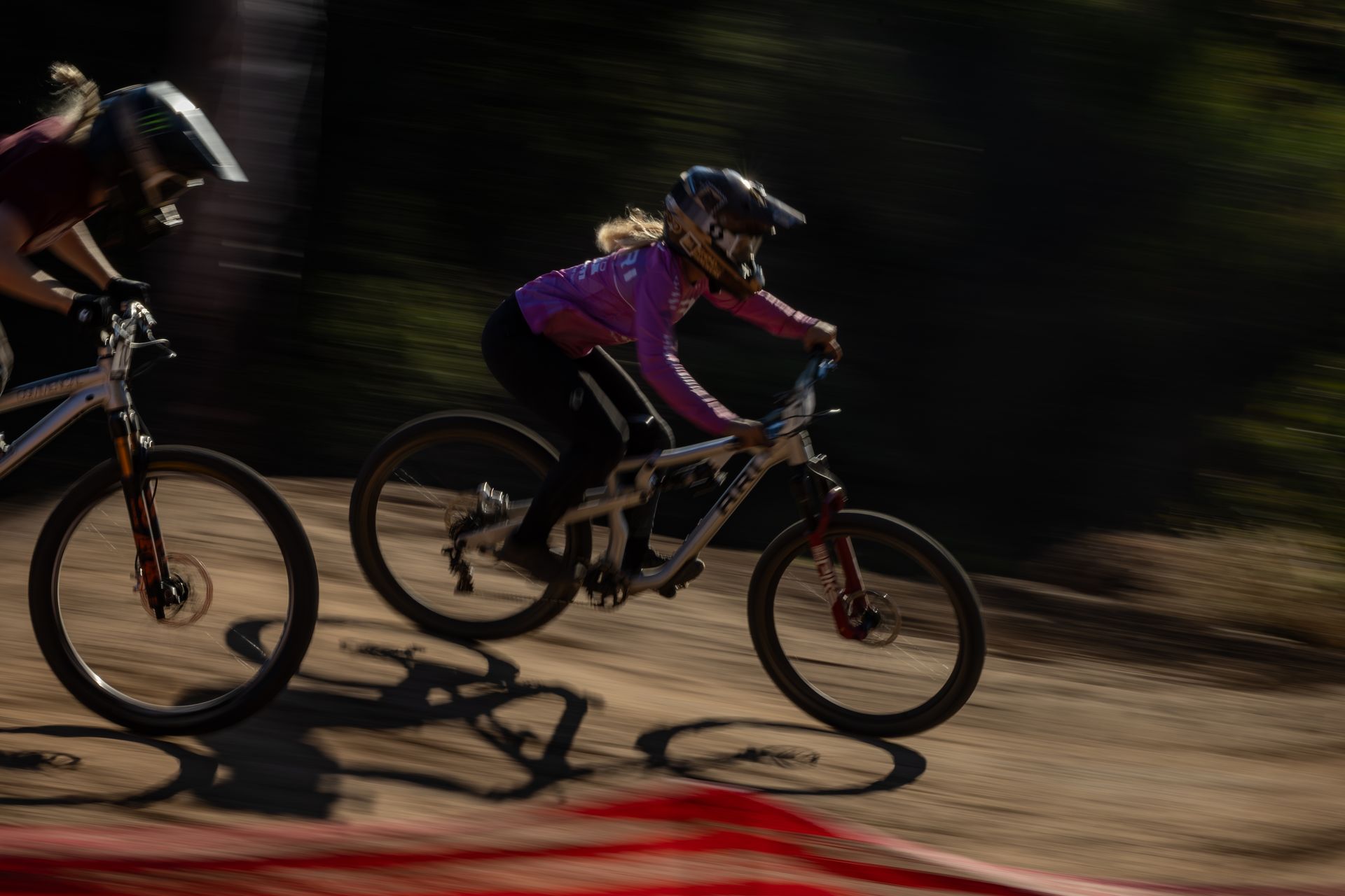 Two mountain bikers speeding down a dirt track, the woman in a pink top and helmet in the foreground.