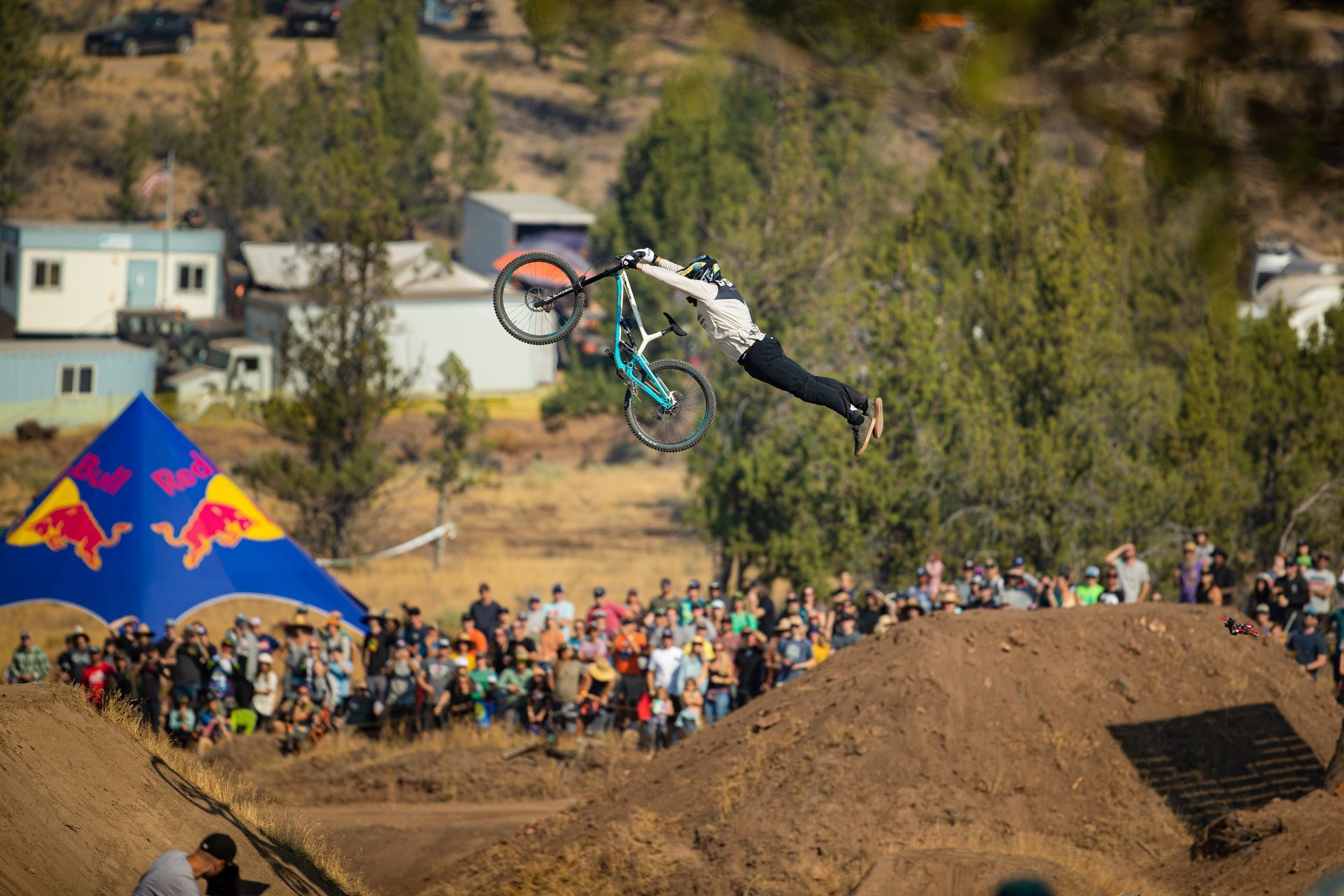 Mountain biker performing a trick in mid-air over a jump, with a crowd and Red Bull tent in the background.