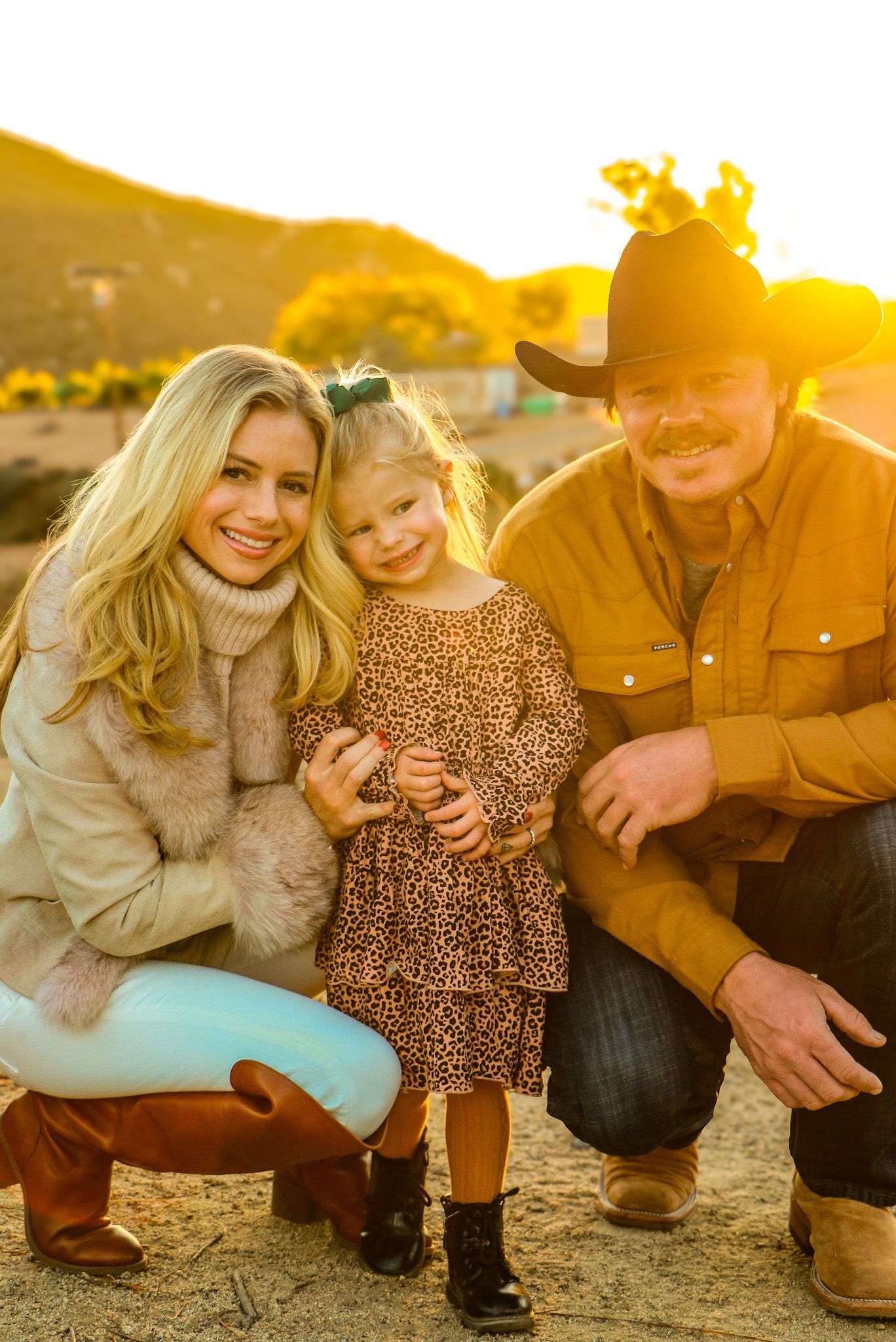 A family poses outdoors in sunset: blonde woman in tan sweater, young girl in dress, and man in cowboy hat.