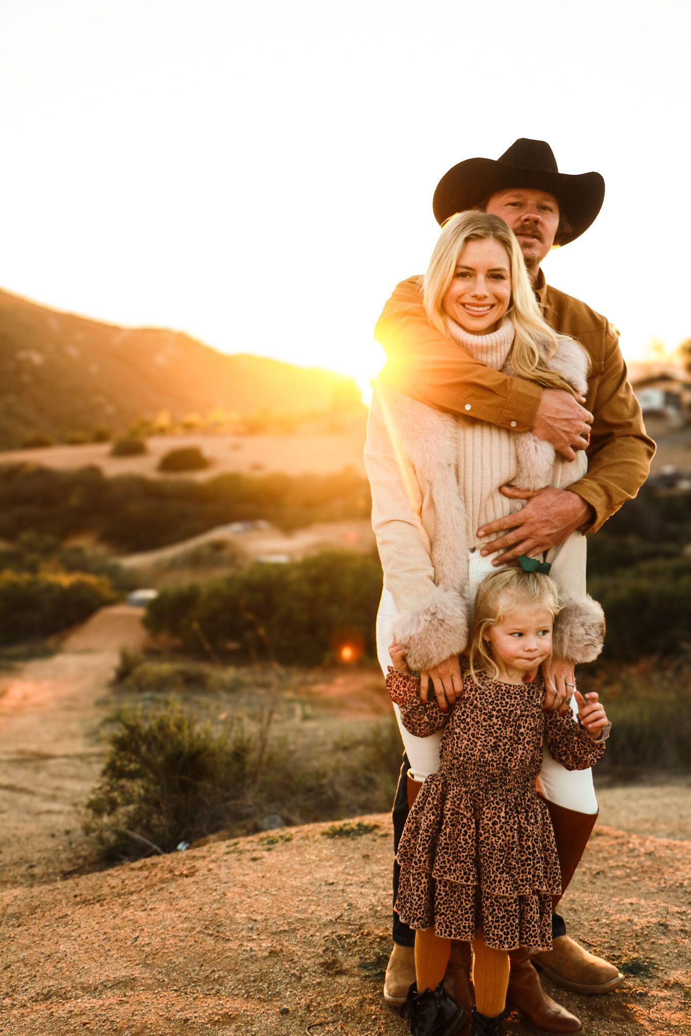 Family of three, embracing outdoors at sunset. Father in cowboy hat, mother in white fur coat
