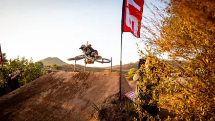 Mountain biker in mid-air over a dirt jump, arms and legs extended. Outdoors, sunny setting, near red and white flags.