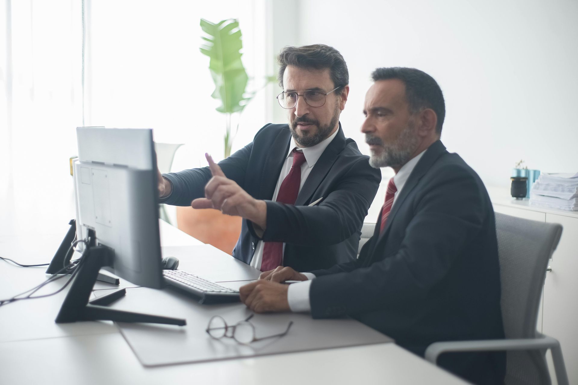 Two men in suits and ties are sitting at a desk looking at a computer screen.