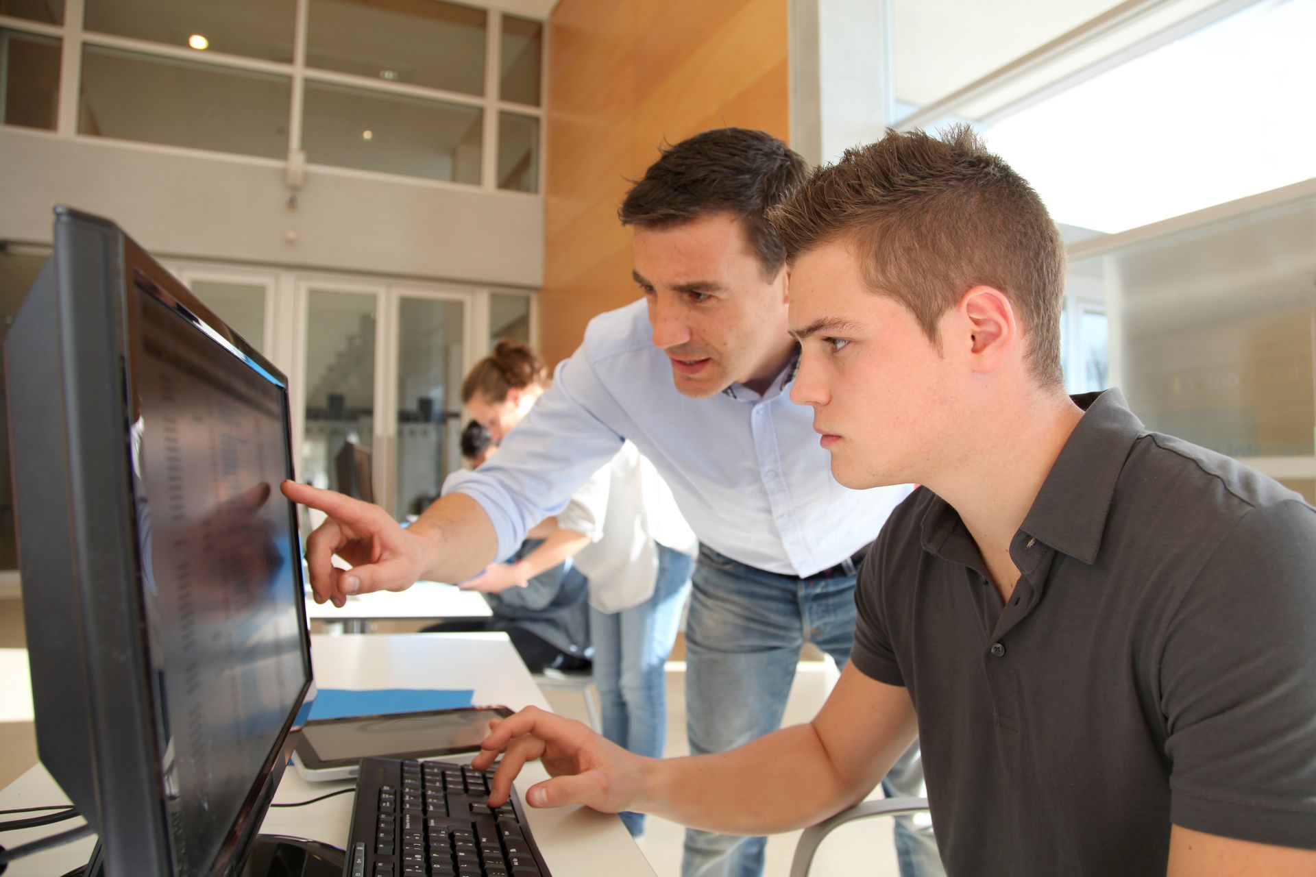 A man is pointing at a computer screen while another man looks on.