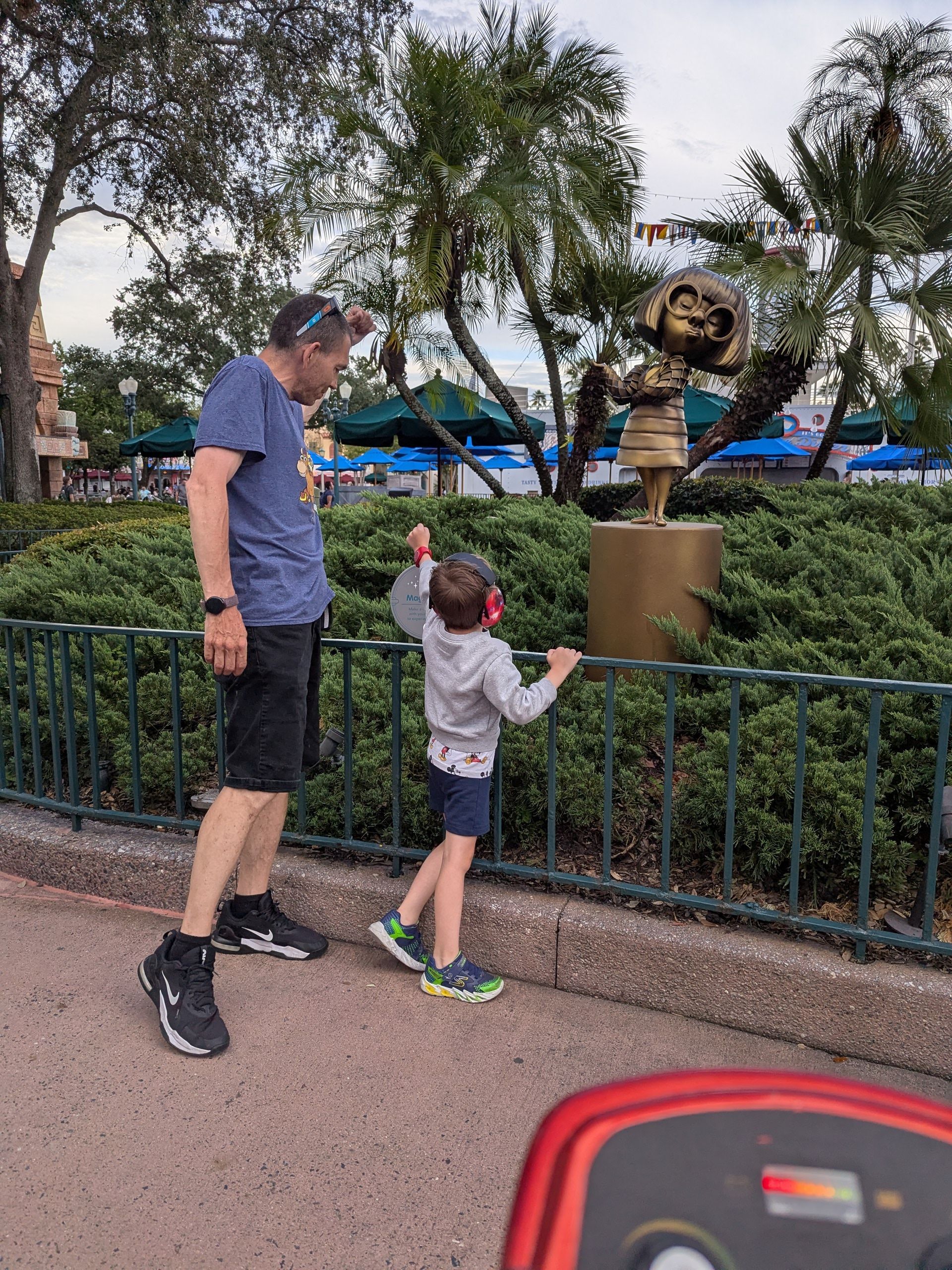 Man and child looking at a golden statue of a girl in front of green bushes.