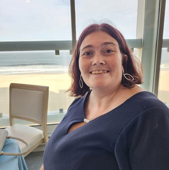 Woman with red hair smiles, wearing a blue dress, by a window with a beach view.