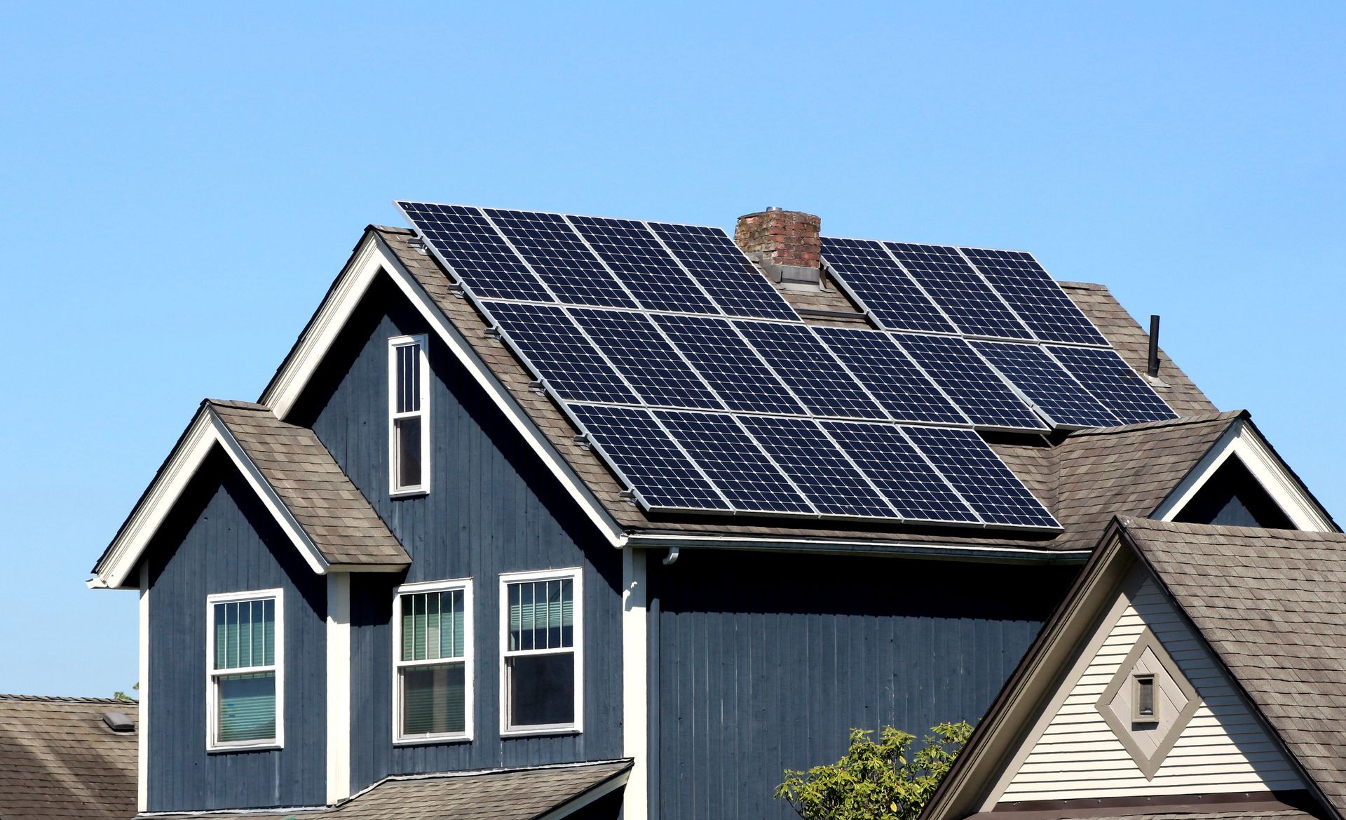 A blue, two-story house with dark shingled roofing and a large solar panel array on the roof against a clear blue sky.