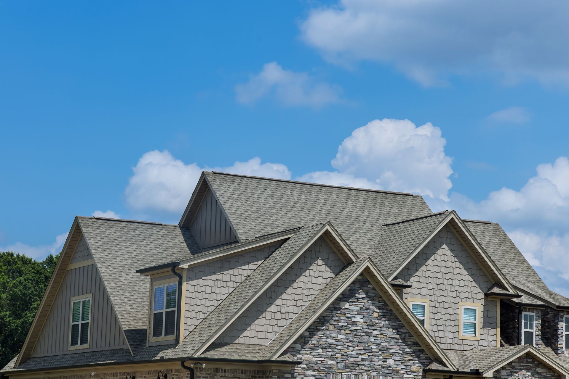 Gabled roof of a home with light gray stone siding and brown roof shingles against a blue sky with white clouds.