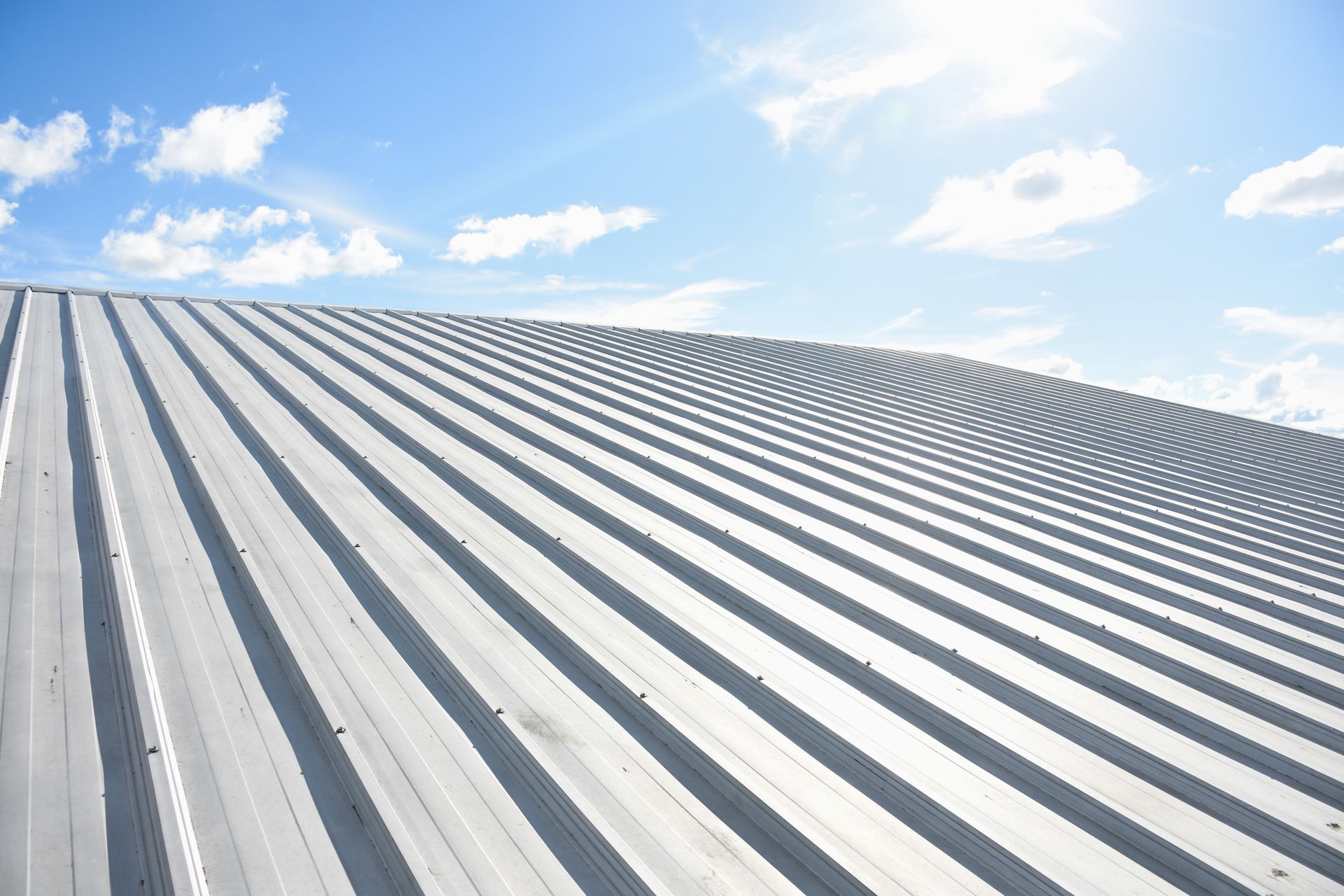 A low-angle view of a silver, corrugated metal roof against a bright blue, partly cloudy sky.