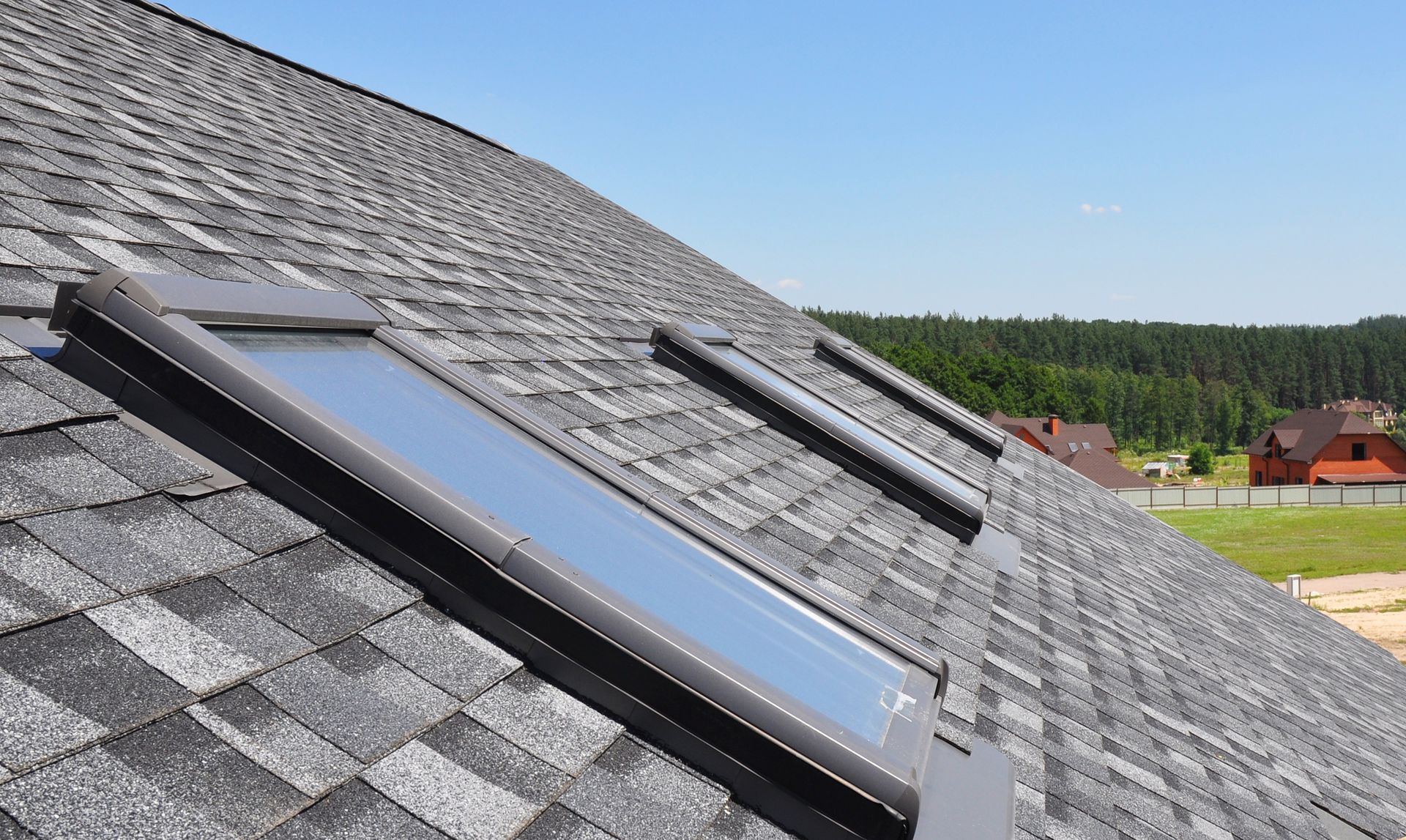 Two skylights installed on a gray asphalt-shingled roof under a clear blue sky.