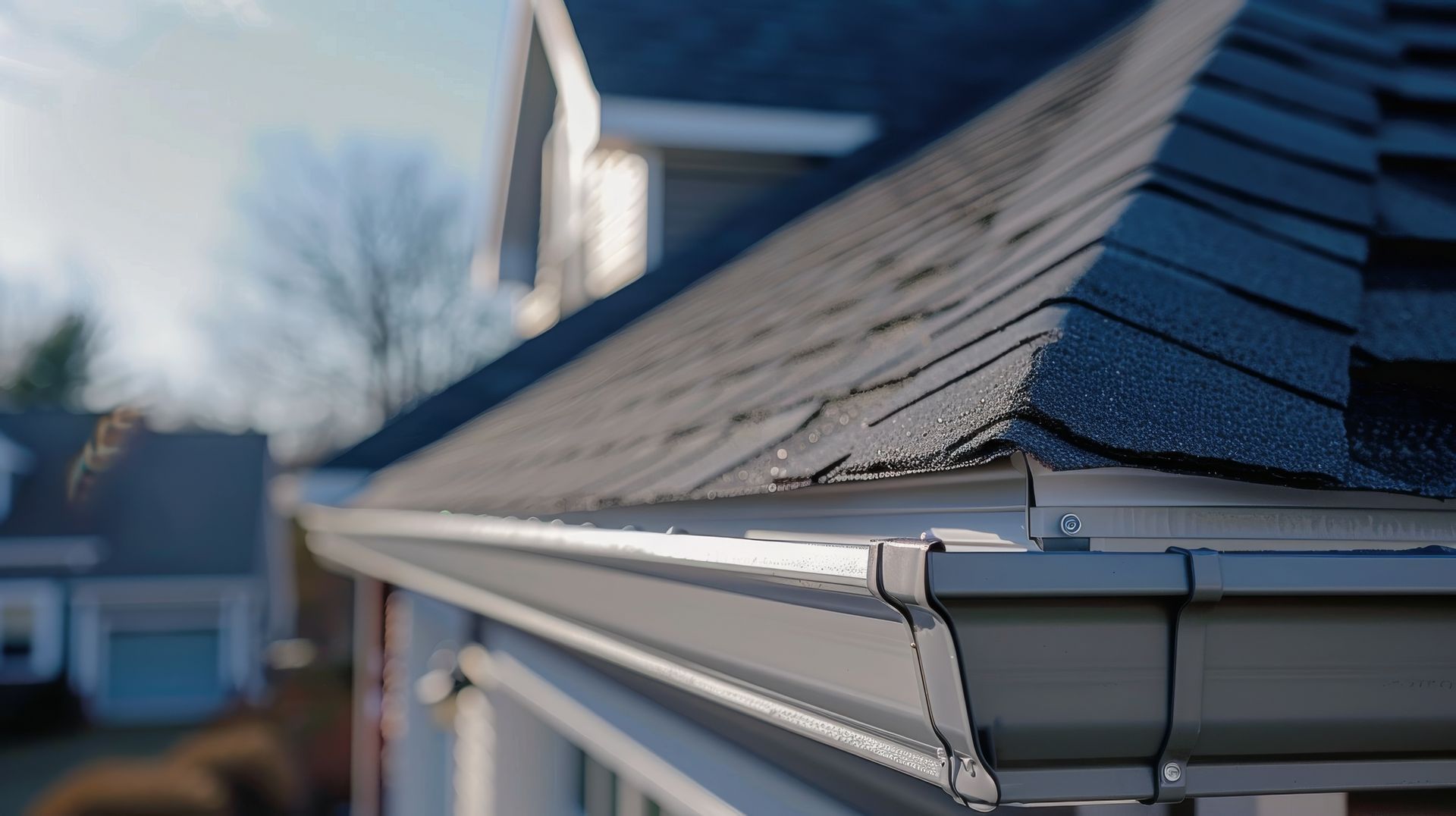 A close-up view of dark grey asphalt roof shingles overlapping the edge of a light-colored metal gutter on a house exterior.
