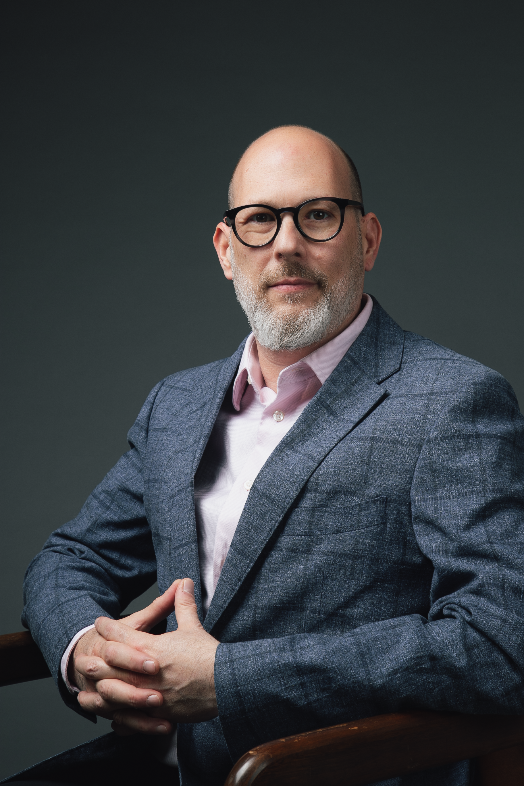 Middle-aged man with glasses, gray beard, in blazer, and pink shirt, sits with hands clasped, looking forward.