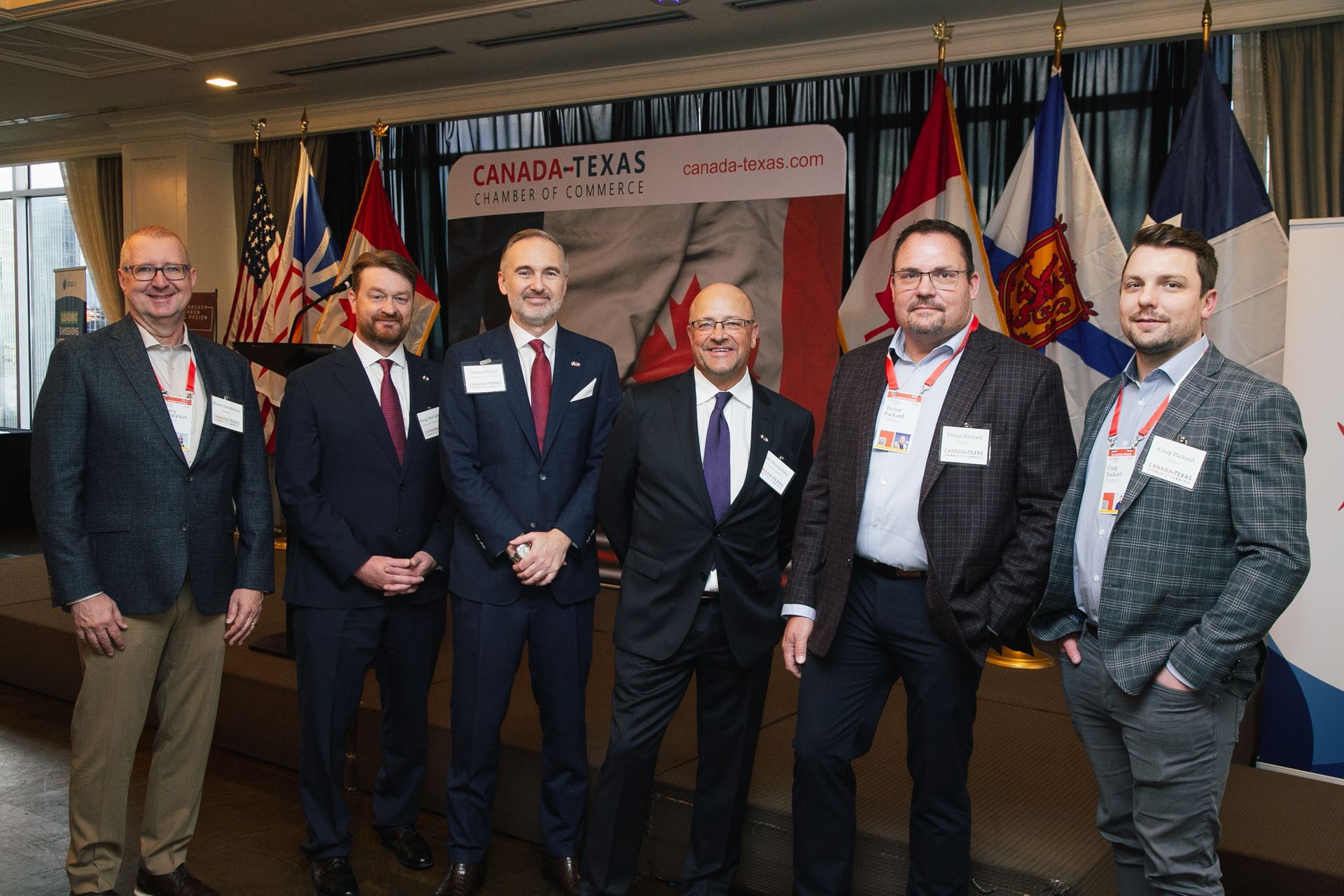 Six people stand in a line on a stage in front of flags and a Canada-Texas business banner at an indoor event.
