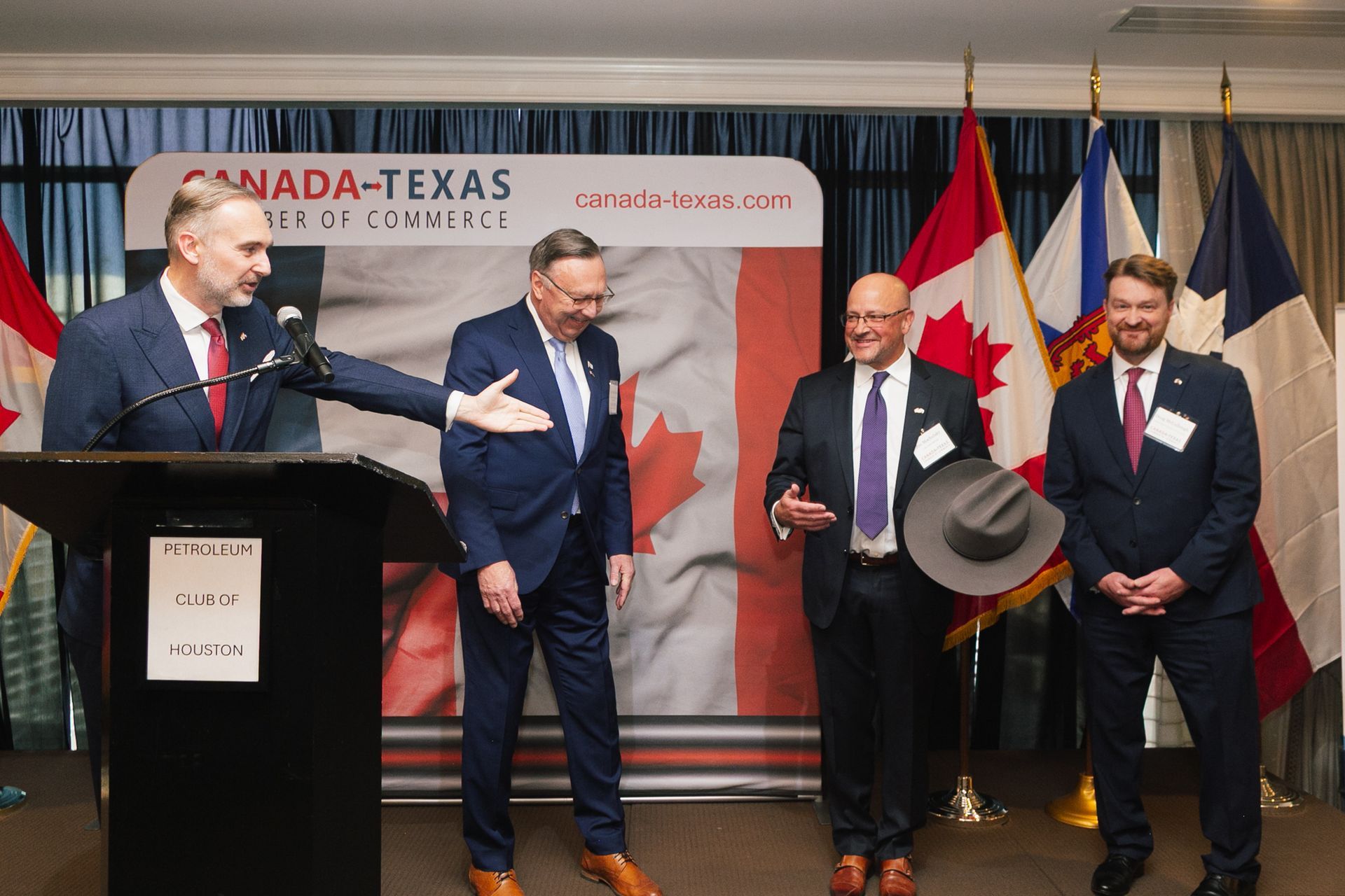 Four men in suits stand before a Canada-Texas Chamber of Commerce banner; one speaks at a podium while another holds a hat.