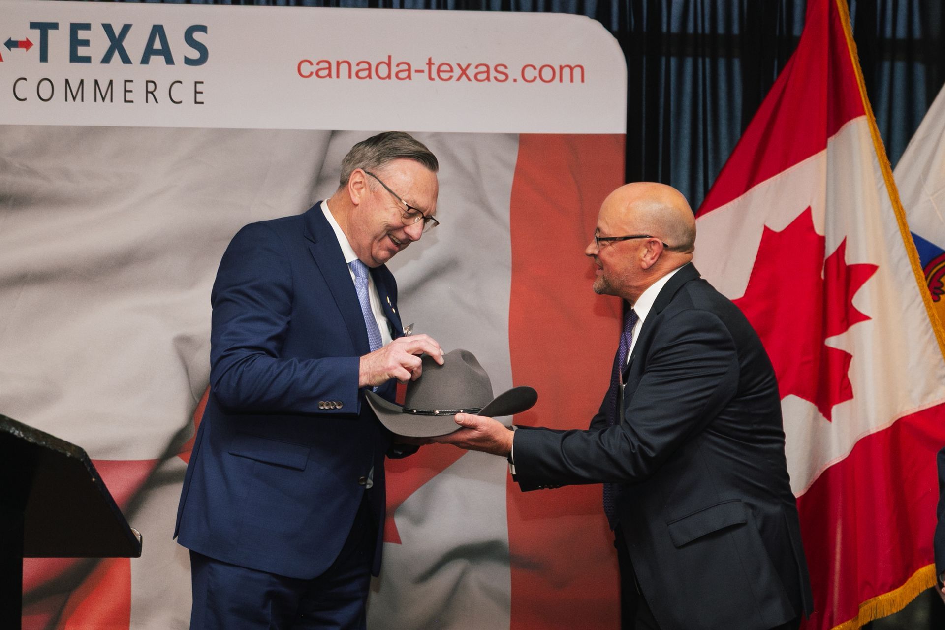 Two men in suits exchange a grey cowboy hat in front of a Canada-Texas Commerce backdrop and a Canadian flag.
