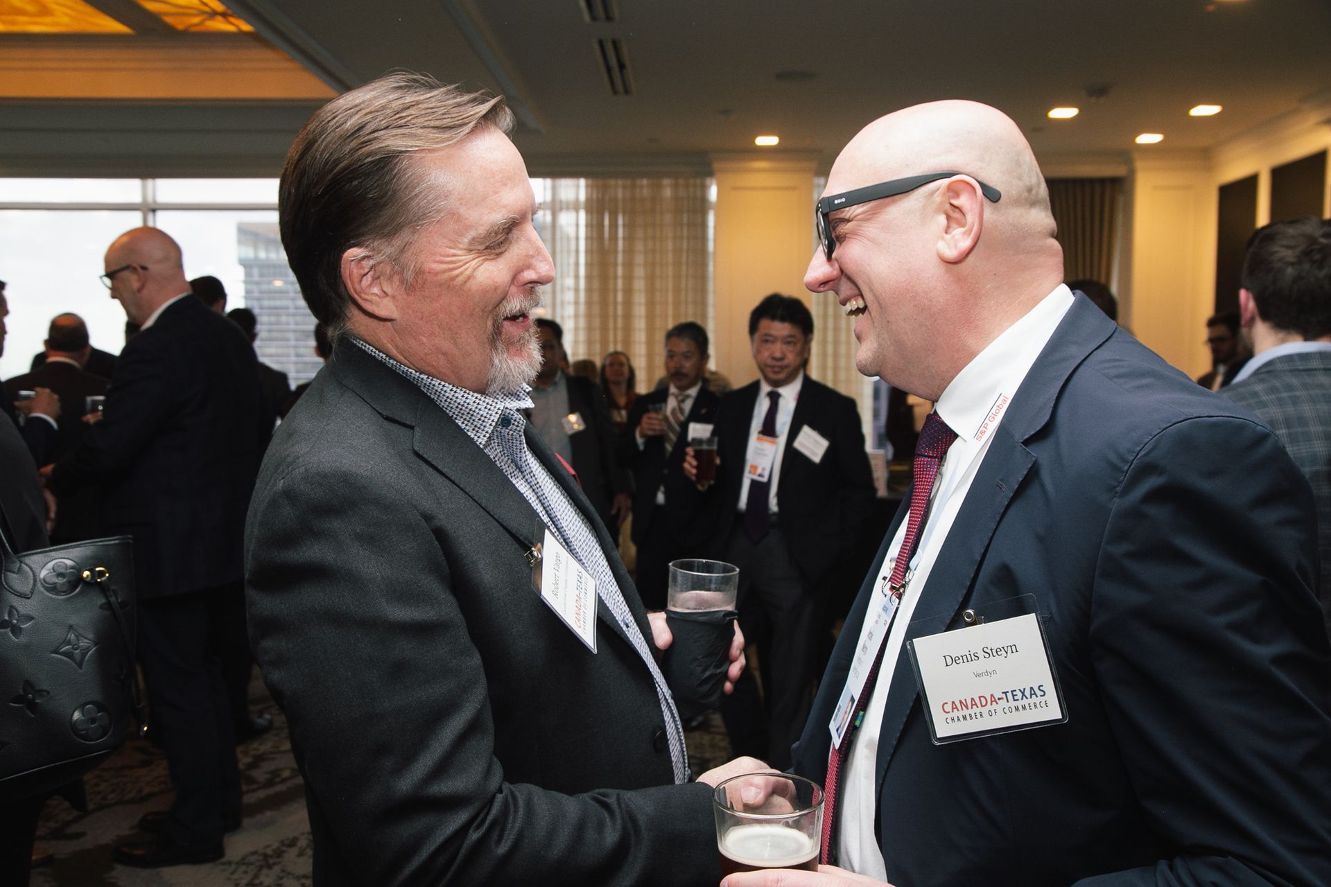 Two men in suits laugh and converse at a brightly lit networking event.