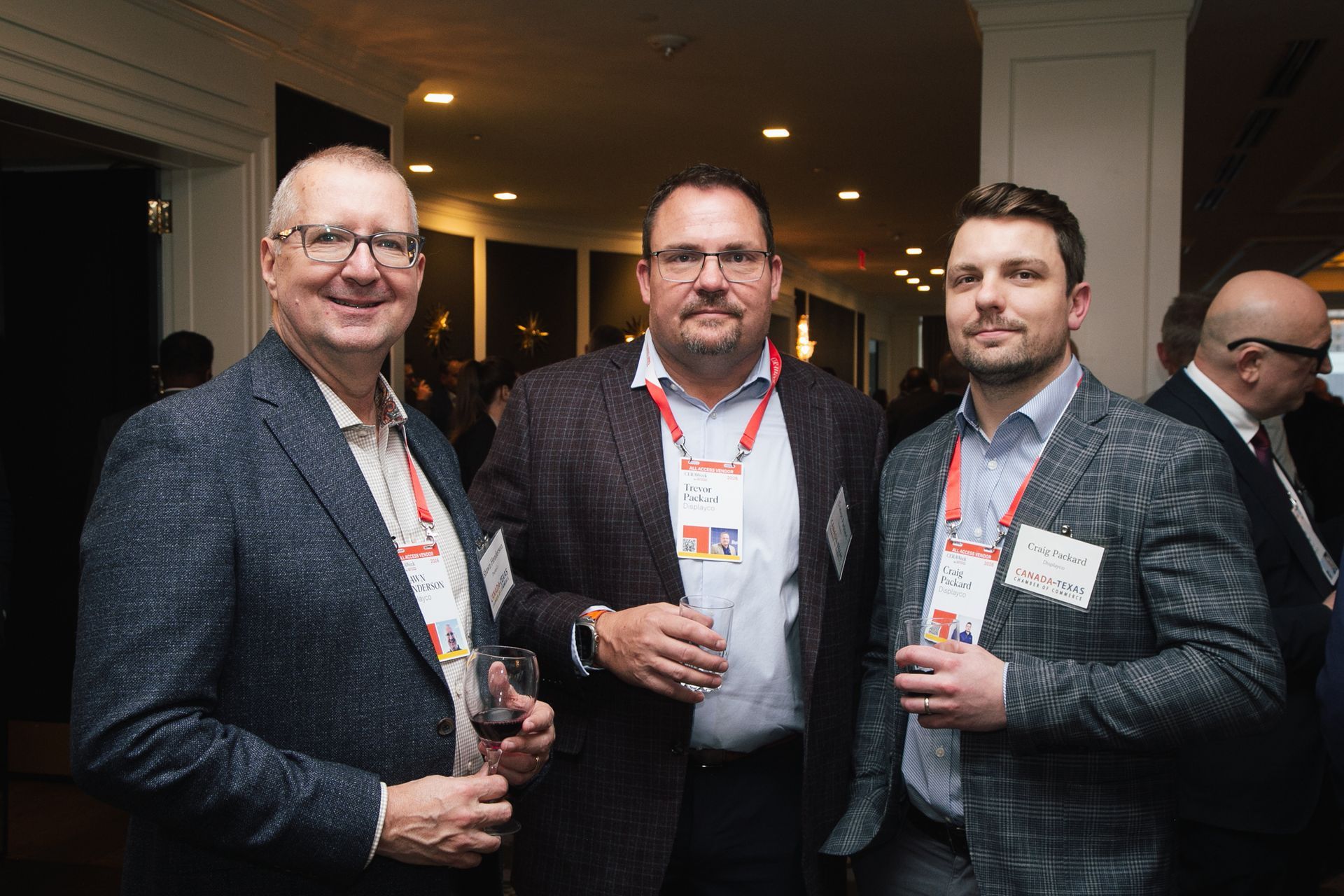 Three people wearing business-casual attire and conference name badges stand in a dimly lit room, holding wine glasses.