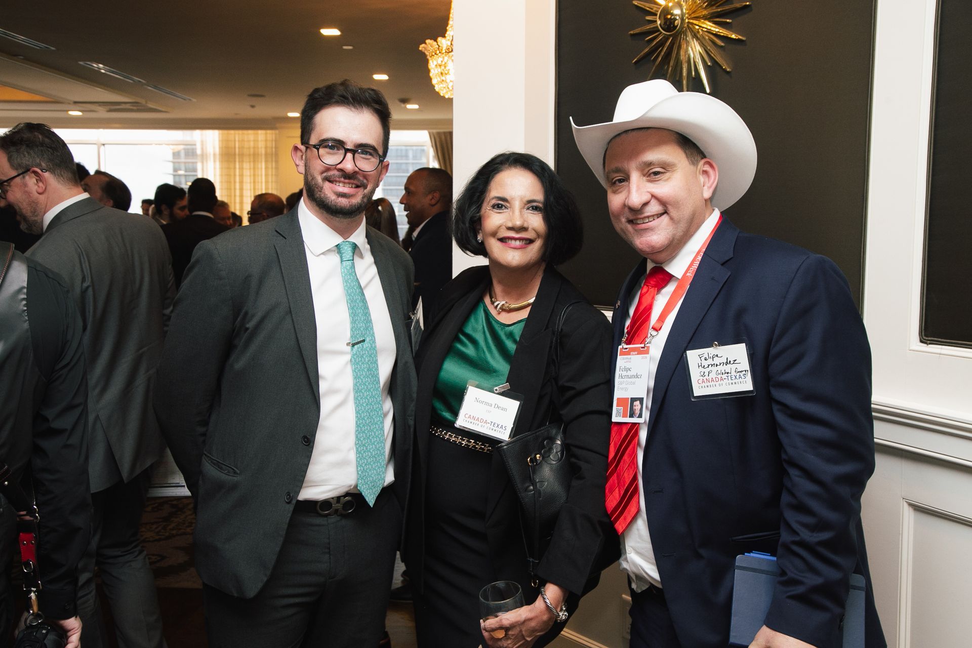 Three people stand together at an indoor event, smiling at the camera. The man on the right wears a cowboy hat.