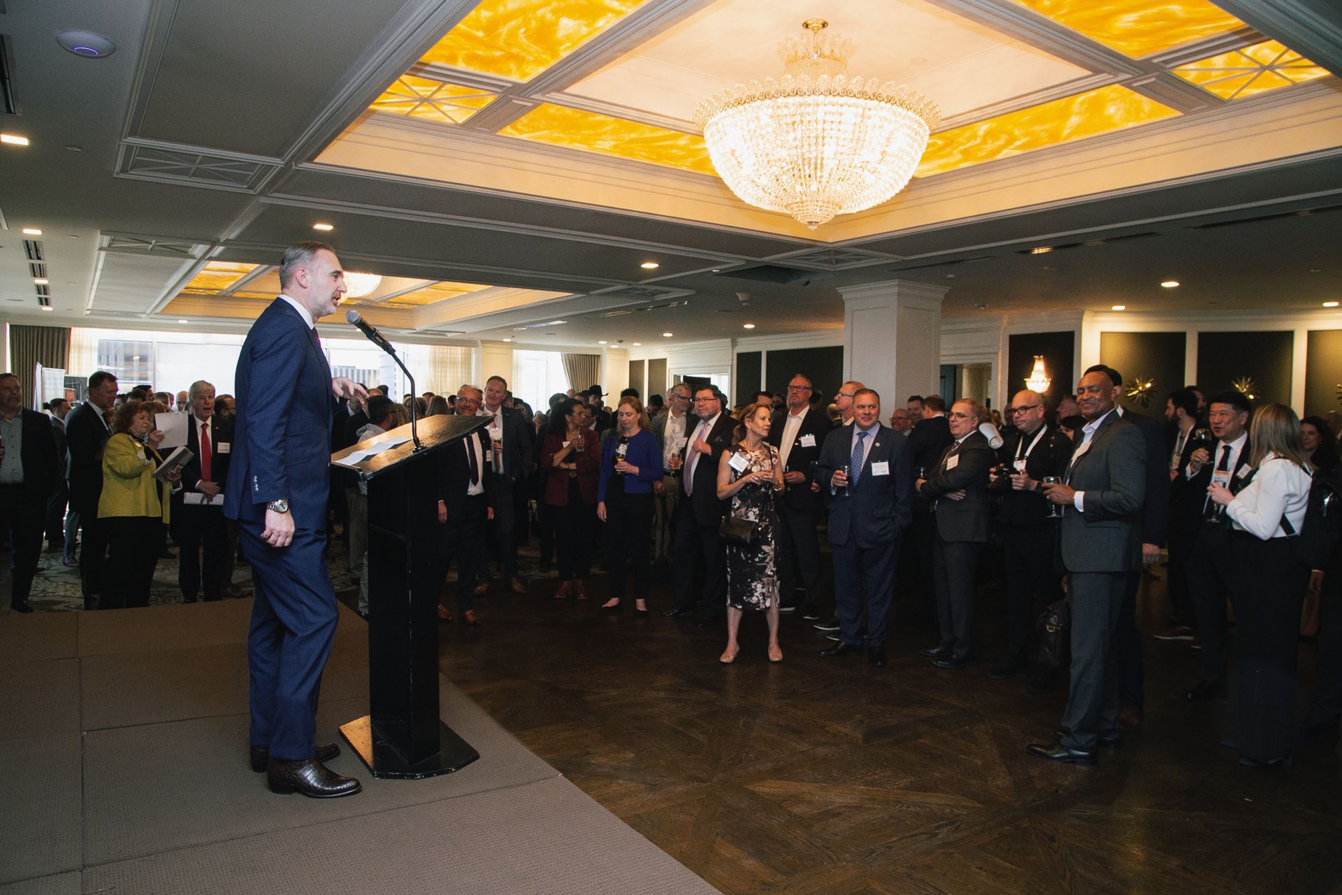 A man in a suit speaks from a podium at a formal indoor event attended by a large group of people in an elegant room.