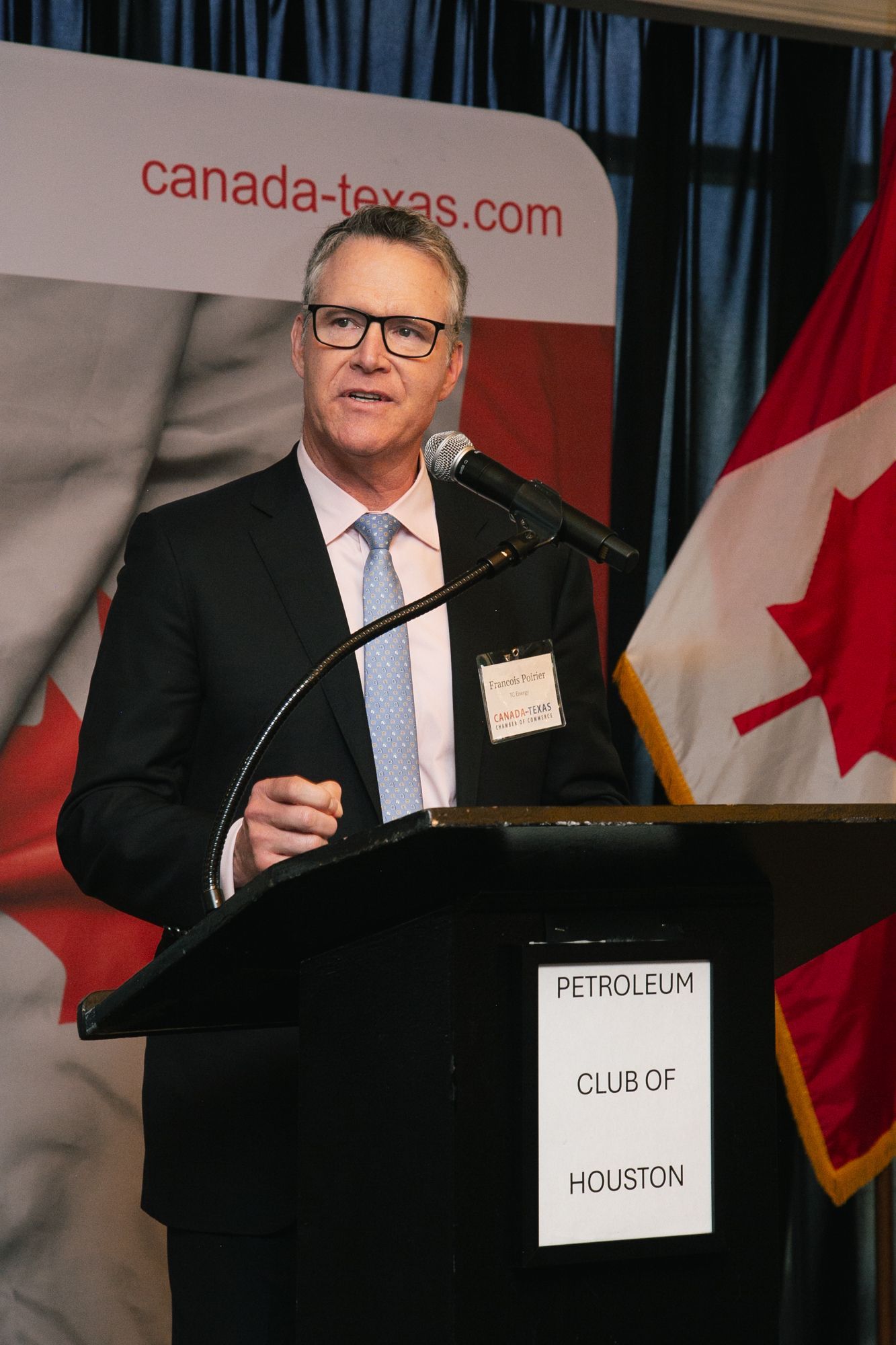 A man in a suit and glasses speaks at a podium with a Canadian flag in the background at the Petroleum Club of Houston.